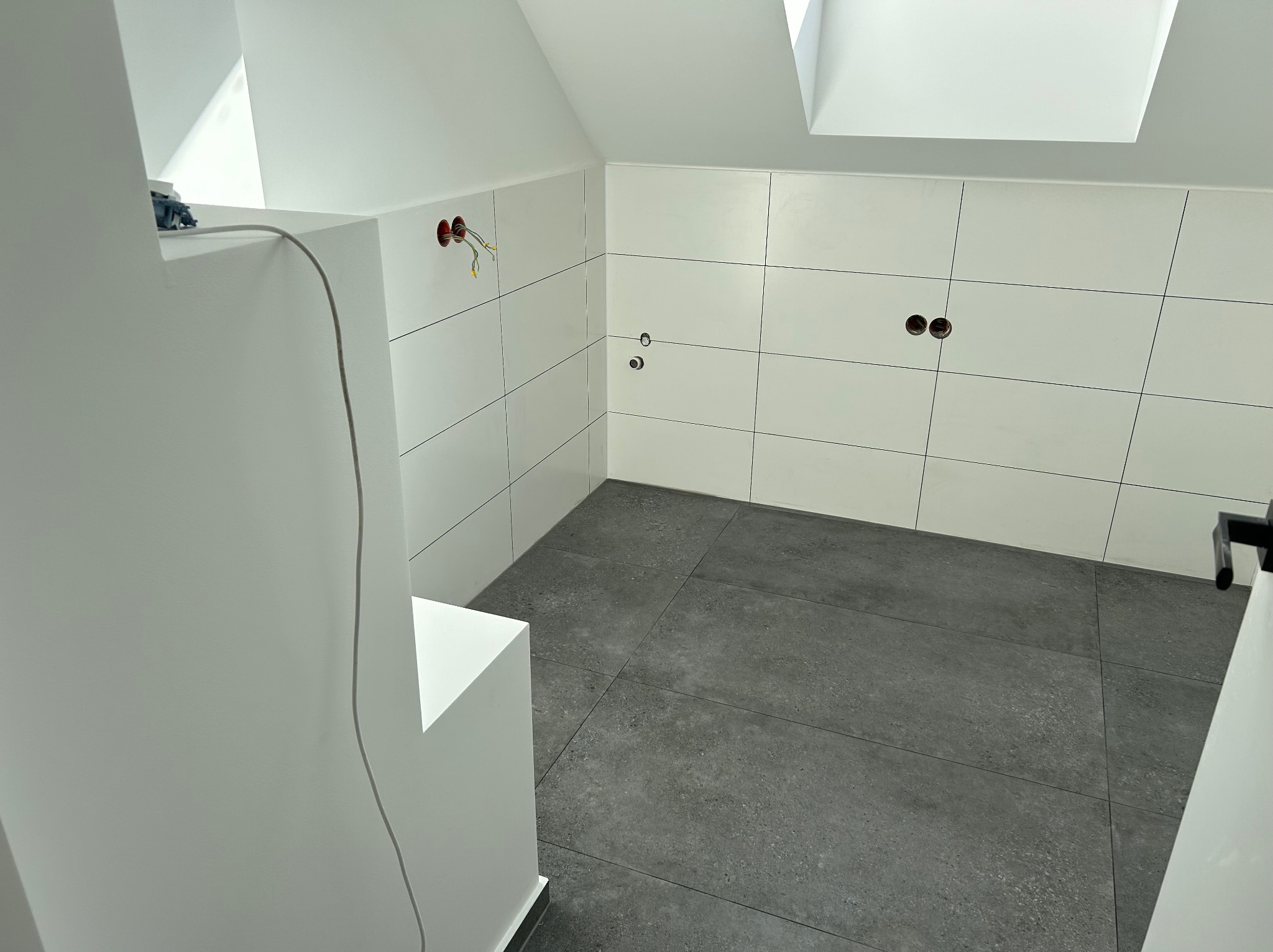 Wnętrze łazienki w trakcie prac wykończeniowych: białe płytki ścienne, szare płytki podłogowe, widoczne kable elektryczne wystające ze ściany, okno dachowe w suficie, minimalistyczny styl.