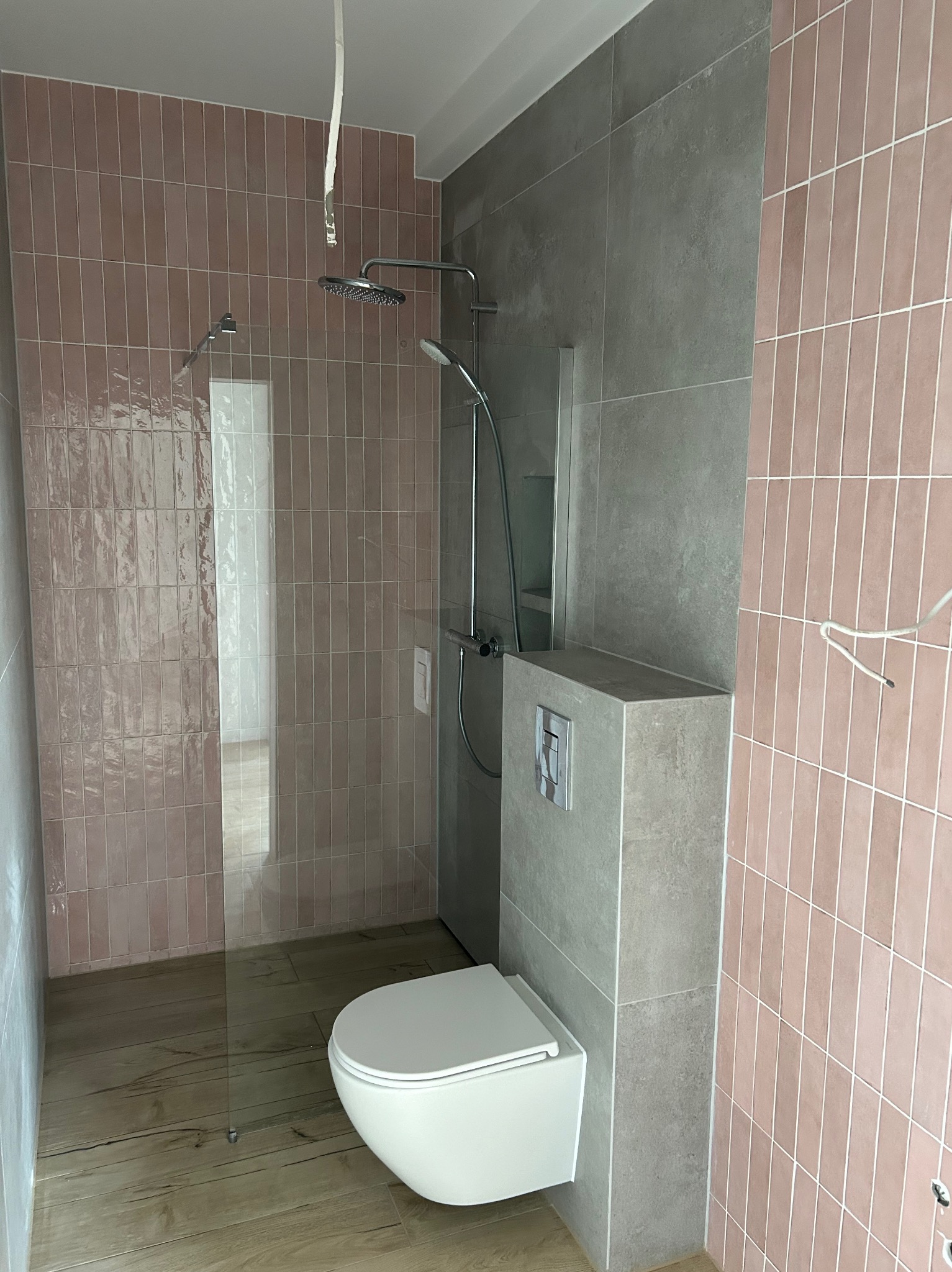 Nowoczesna łazienka z różowymi, pionowo ułożonymi płytkami, szarymi płytami wielkoformatowymi, drewnopodobną podłogą, szklanym panelem prysznicowym i wiszącą toaletą z zabudowaną spłuczką.