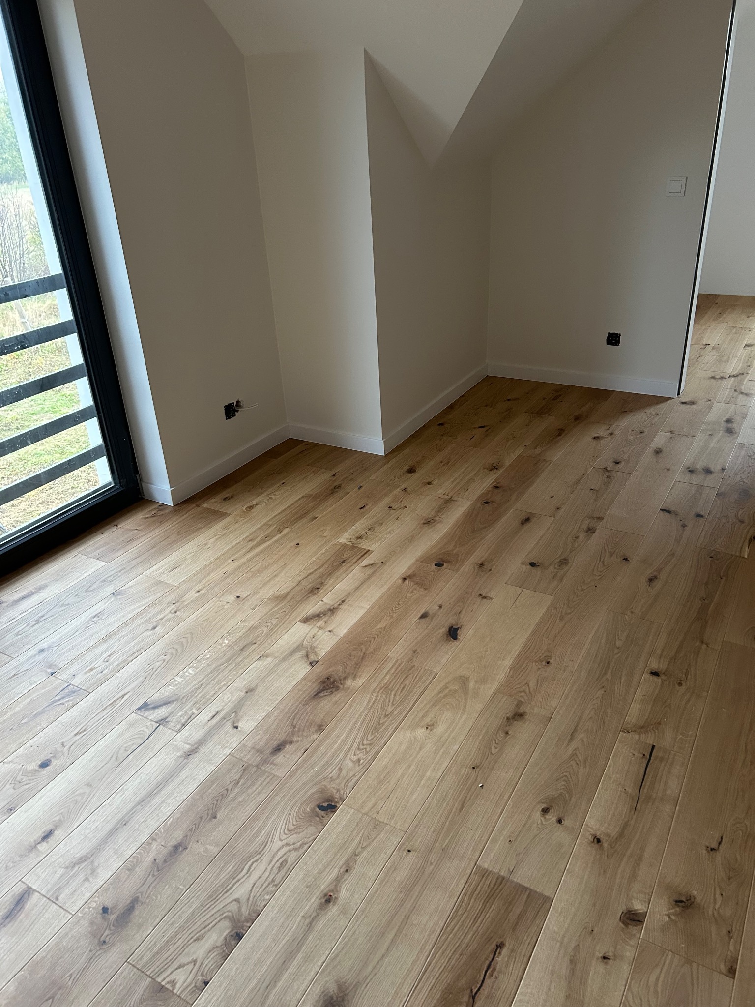 Jasne, drewniane panele podłogowe ułożone w nowym pokoju z oknem balkonowym i świeżo pomalowanymi ścianami w odcieniach bieli.