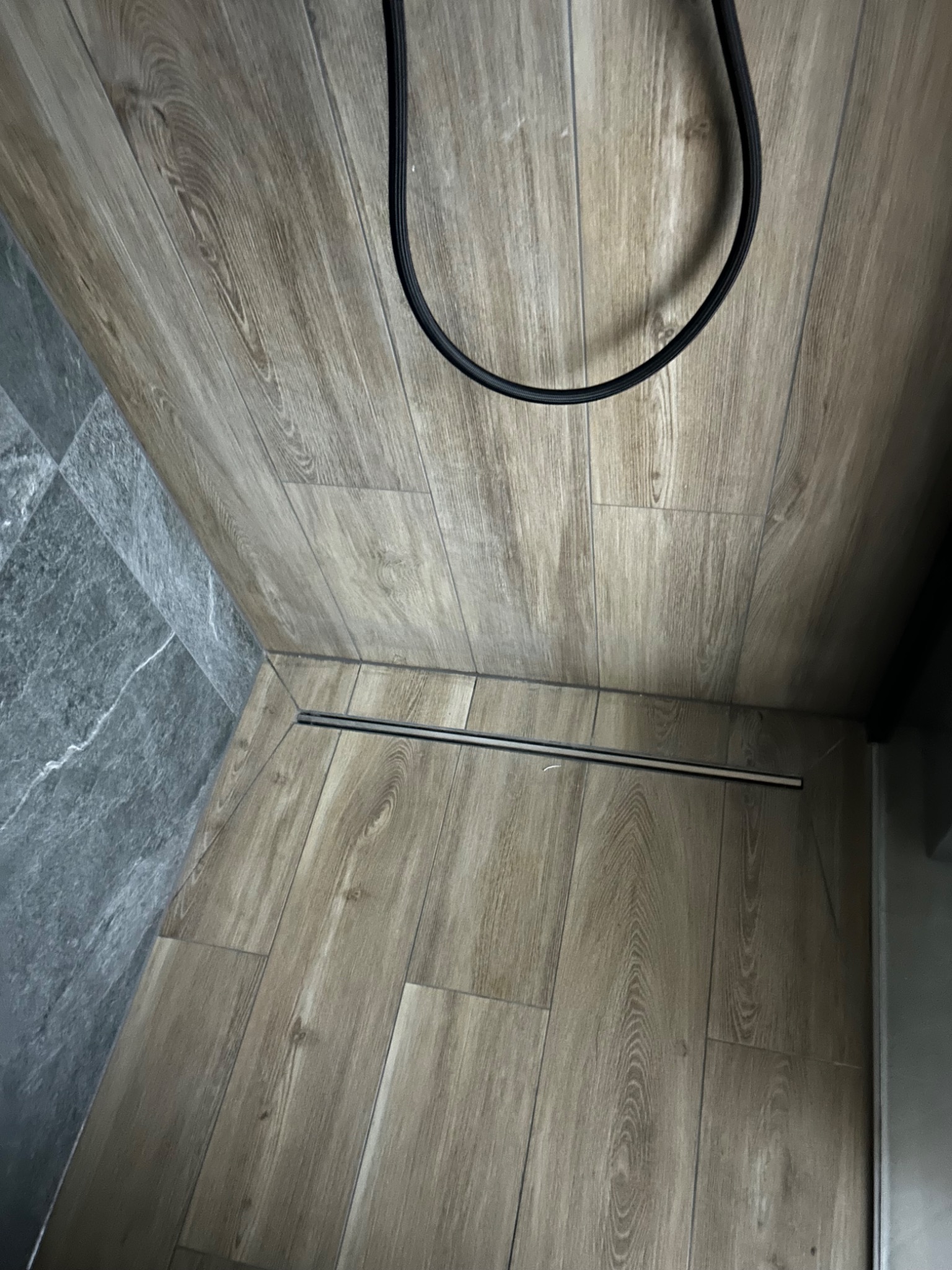 Szczegół wykończonej podłogi w łazience z odpływem liniowym, panelami imitującymi drewno i fragmentem ściany wyłożonej szarą płytką imitującą kamień.