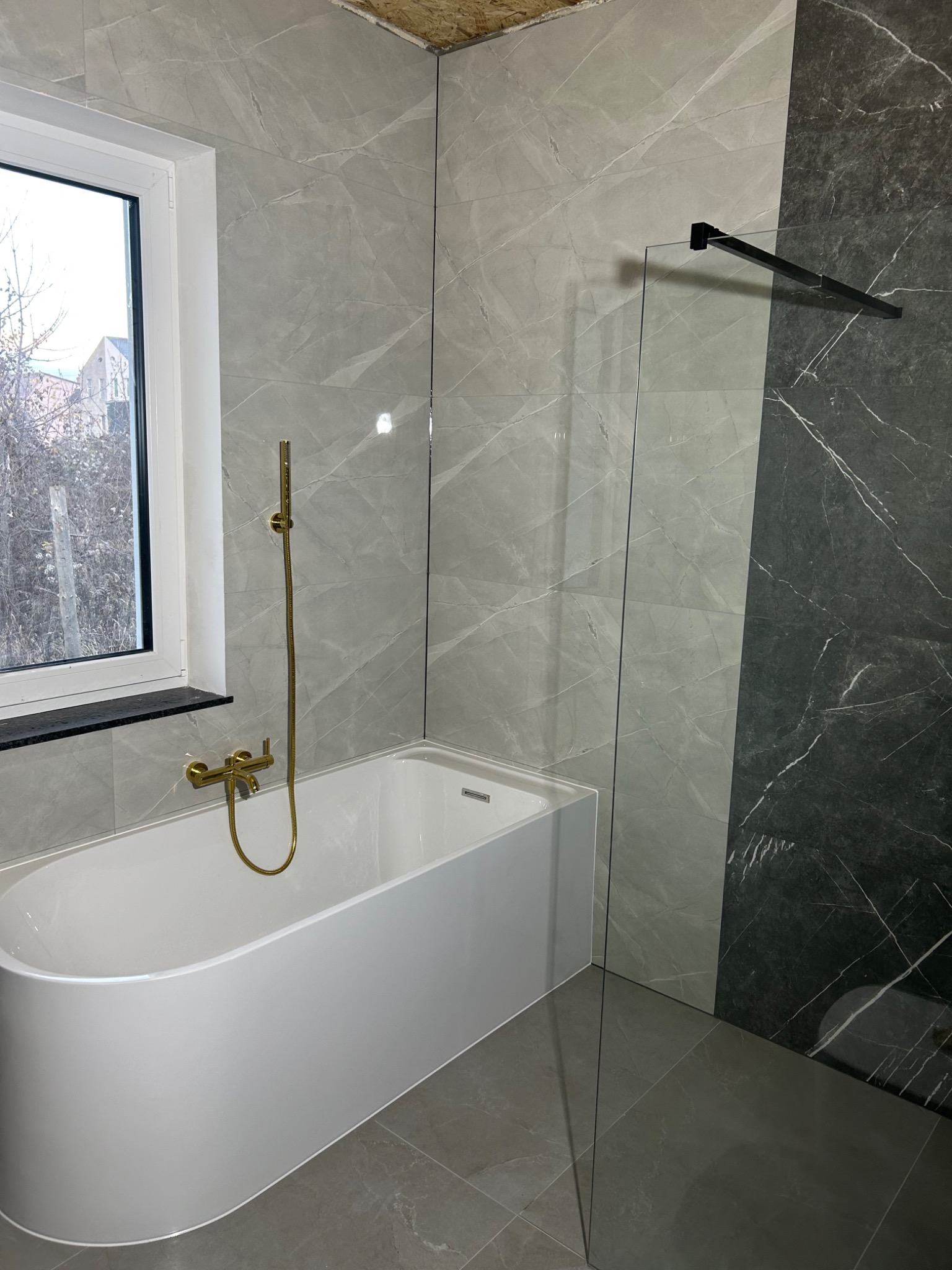 Nowoczesna łazienka z wanną, złotą armaturą i szarymi płytkami imitującymi marmur, oddzielona szklanym panelem z czarnym uchwytem.