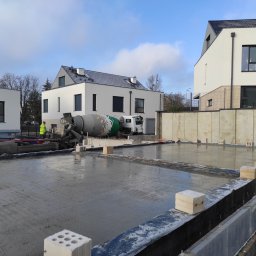 Andrii Vuichyk - Wylany beton na przygotowany teren pod budowę domu, widoczny mikser do betonu, pracownik w żółtej kamizelce oraz w tle nowoczesne budynki.