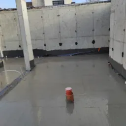 Świeżo wylana betonowa posadzka wewnątrz budynku z surowymi, betonowymi ścianami i zaizolowanymi fundamentami, widoczne rury wentylacyjne i elementy szalunku.