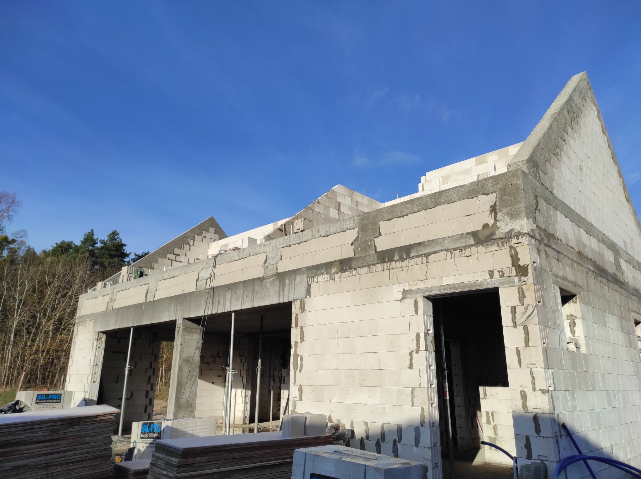 Wznoszenie ścian parterowego domu z betonu komórkowego, widoczne zbrojenie i podpory stropu, na pierwszym planie palety z bloczkami.