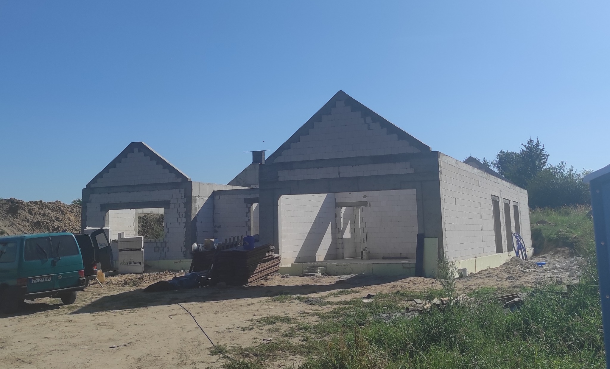 Wznoszenie ścian parterowego domu z betonu komórkowego, widoczne ściany zewnętrzne i działowe, zarys dachu, izolacja fundamentów styropianem, w tle składowany materiał budowlany i samochód dostawczy.