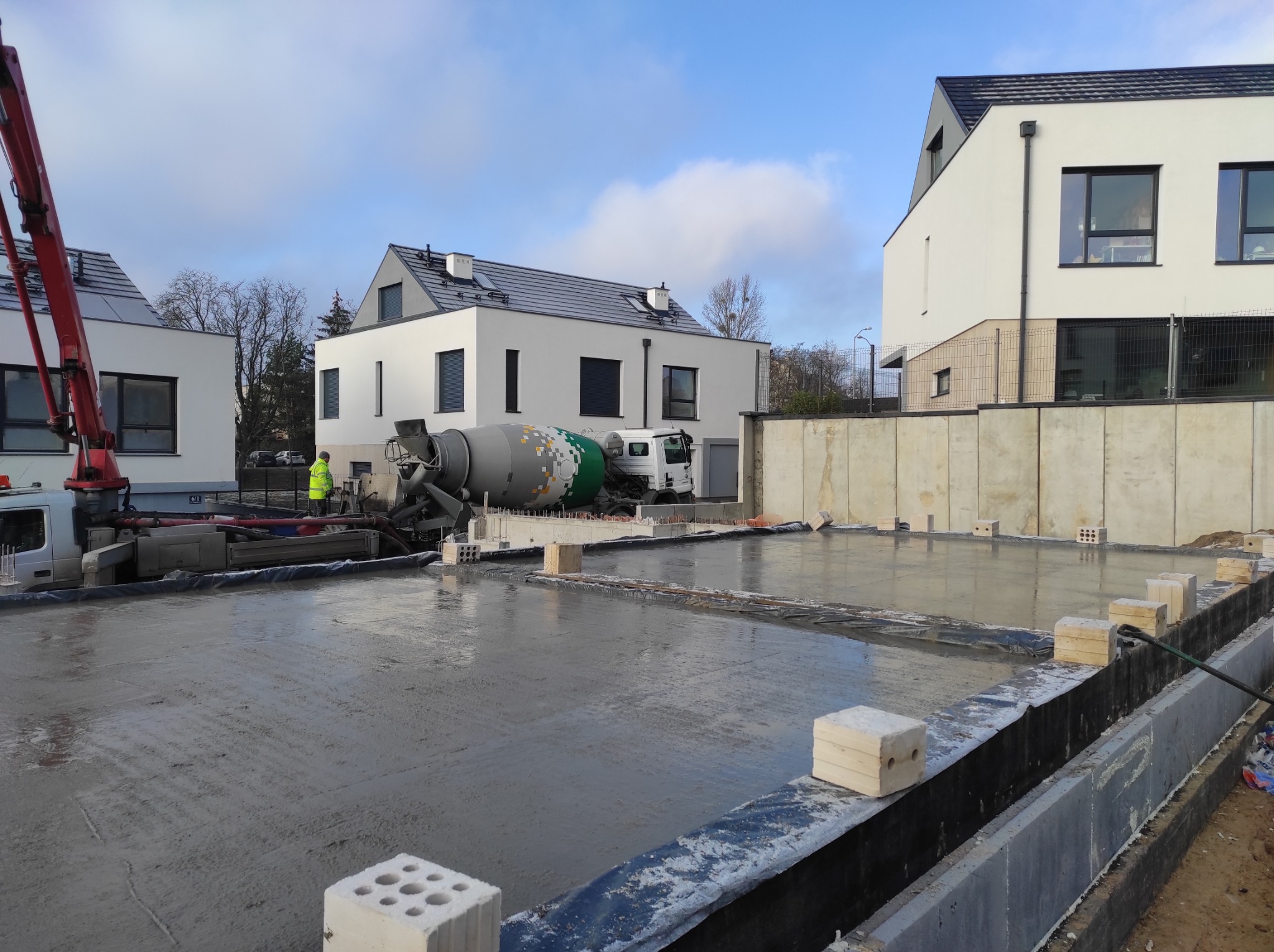 Wylany beton na przygotowany teren pod budowę domu, widoczny mikser do betonu, pracownik w żółtej kamizelce oraz w tle nowoczesne budynki.