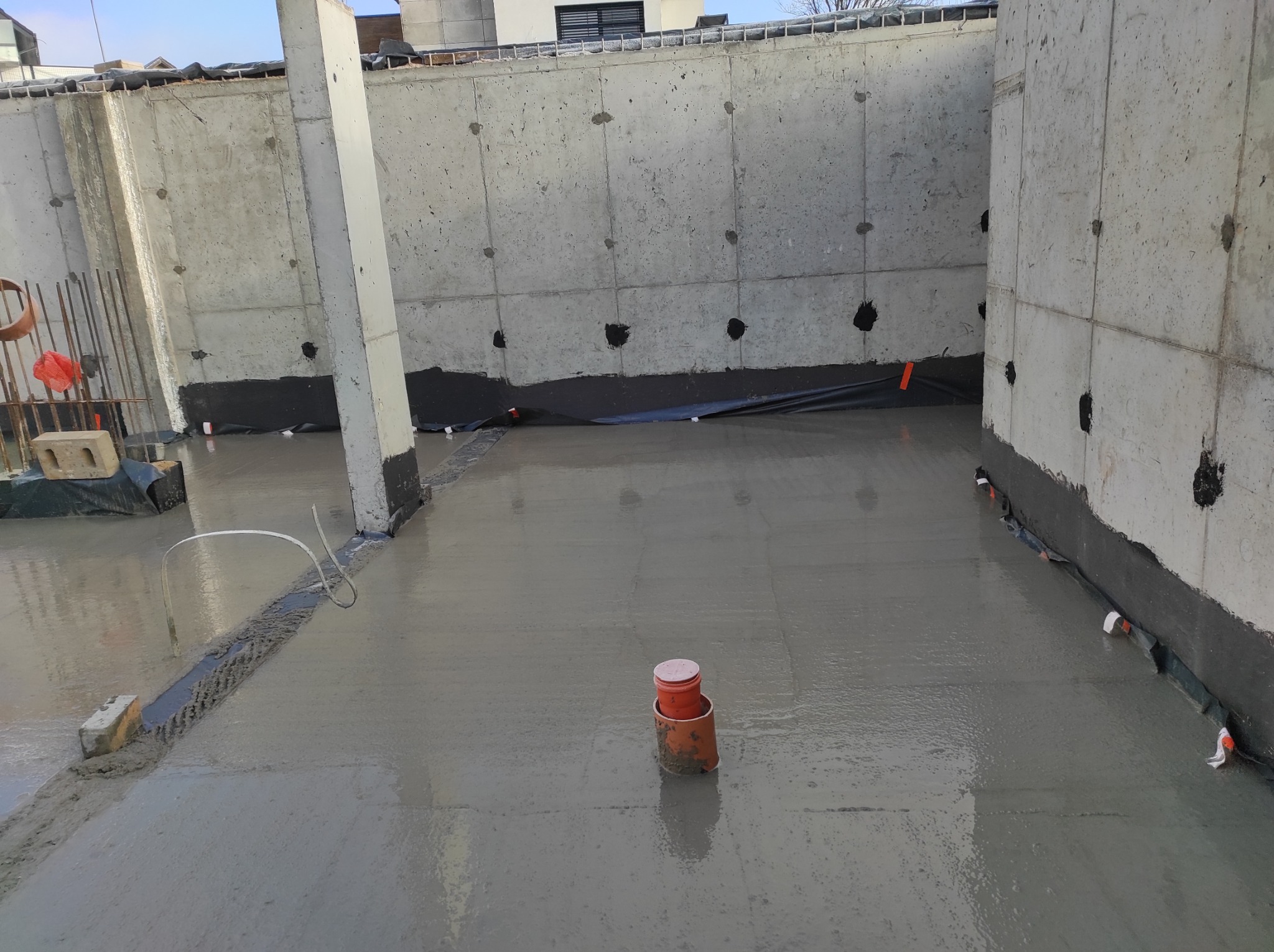 Świeżo wylana betonowa posadzka wewnątrz budynku z surowymi, betonowymi ścianami i zaizolowanymi fundamentami, widoczne rury wentylacyjne i elementy szalunku.