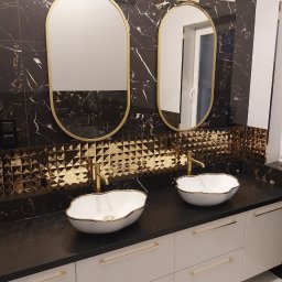 Usługi Remontowe, Wykończenie Wnętrz Artur Kowalski - Elegancka łazienka z dwoma umywalkami w kształcie muszli z złotymi obwódkami, złotymi bateriami, lustrami w złotych ramach i czarnymi płytkami imitującymi marmur ze złotymi żyłami, uzupełniona...