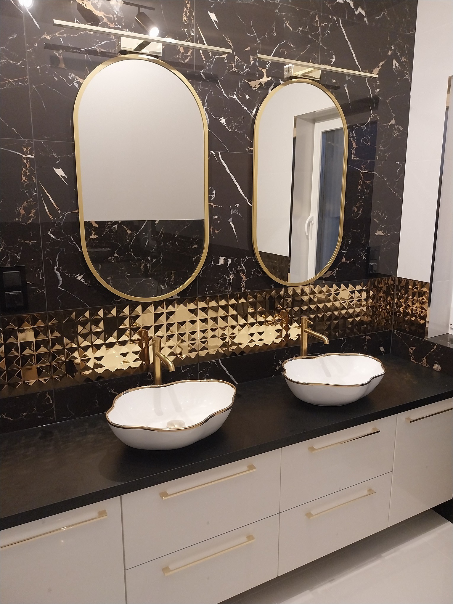 Elegancka łazienka z dwoma umywalkami w kształcie muszli z złotymi obwódkami, złotymi bateriami, lustrami w złotych ramach i czarnymi płytkami imitującymi marmur ze złotymi żyłami, uzupełniona...