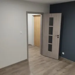 Wnętrze pokoju po remoncie z otwartymi drzwiami wewnętrznymi z szybami, widoczne panele podłogowe w odcieniach beżu i szarości, ściany pomalowane na biało i granatowo, listwy przypodłogowe...