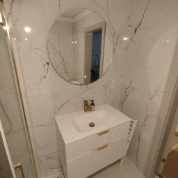 Budmaan - Biała łazienka z marmurowymi płytkami, okrągłym lustrem z podświetleniem i szafką umywalkową z frezowanymi frontami. Złote detale armatury i uchwytów.