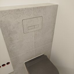 Budmaan - Szary, podtynkowy zestaw toaletowy z płytkami imitującymi beton i ciemnoszarą deską sedesową w minimalistycznej łazience.