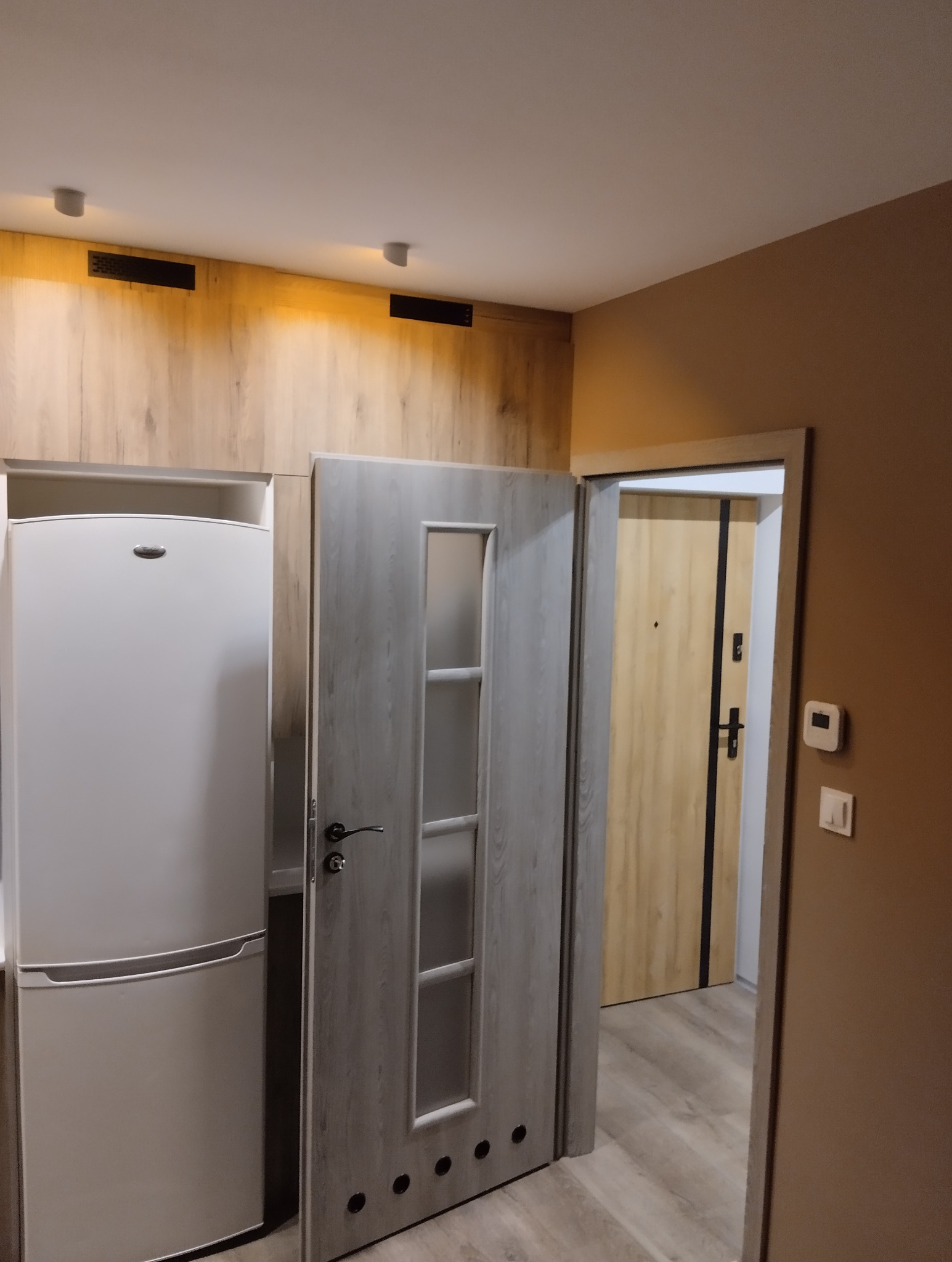Wnętrze mieszkania po remoncie: widok na otwarte, wewnętrzne drzwi z przeszkleniami, obok lodówka w zabudowie i kolejne drzwi w głębi, z drewnopodobnymi panelami na ścianie.