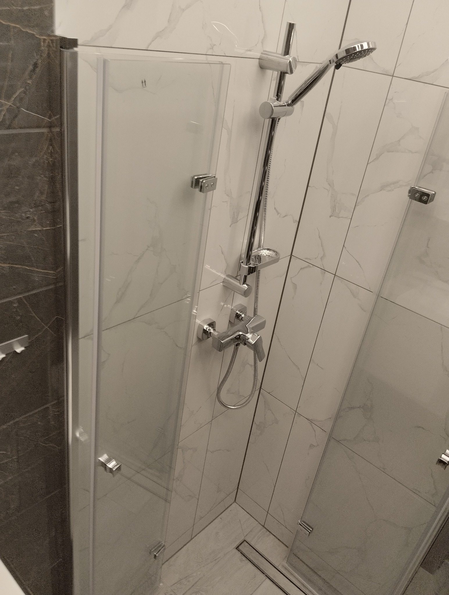 Kabina prysznicowa z chromowaną armaturą i odpływem liniowym, wykończona płytkami imitującymi marmur.