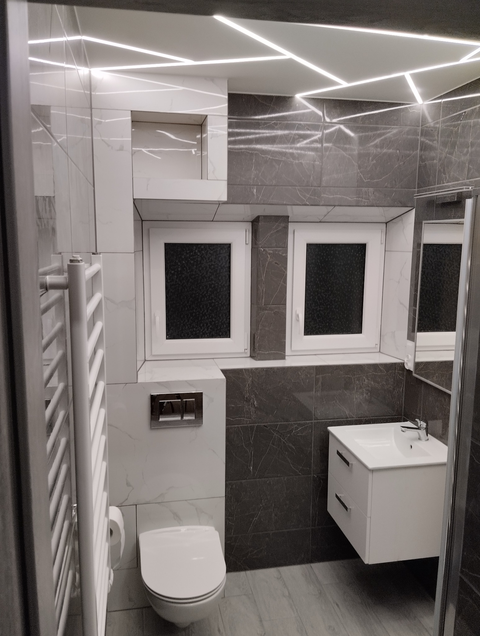 Nowoczesna łazienka w Bytowie z szarymi i białymi płytkami imitującymi marmur, podwieszana toaleta, biała szafka z umywalką i dekoracyjne oświetlenie sufitowe LED.