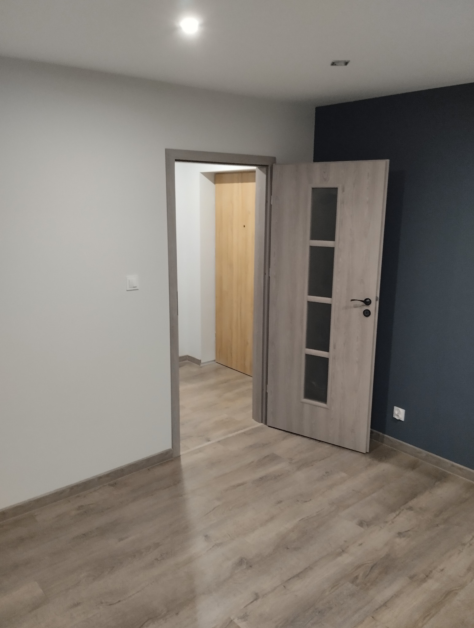 Wnętrze pokoju po remoncie z otwartymi drzwiami wewnętrznymi z szybami, widoczne panele podłogowe w odcieniach beżu i szarości, ściany pomalowane na biało i granatowo, listwy przypodłogowe...