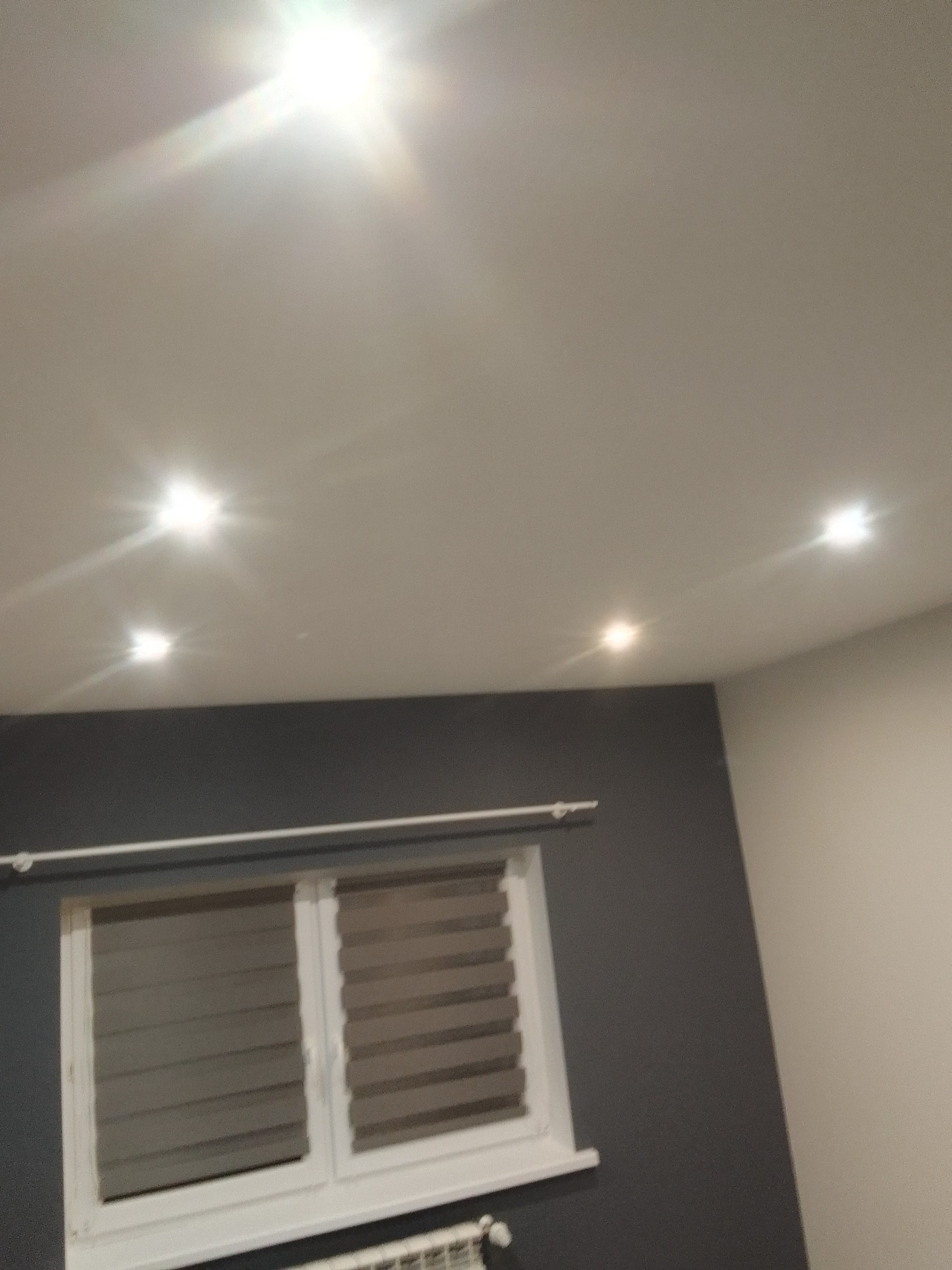 Szare wnętrze z białym oknem z żaluzjami poziomymi, grzejnikiem pod oknem i czterema punktowymi lampami sufitowymi