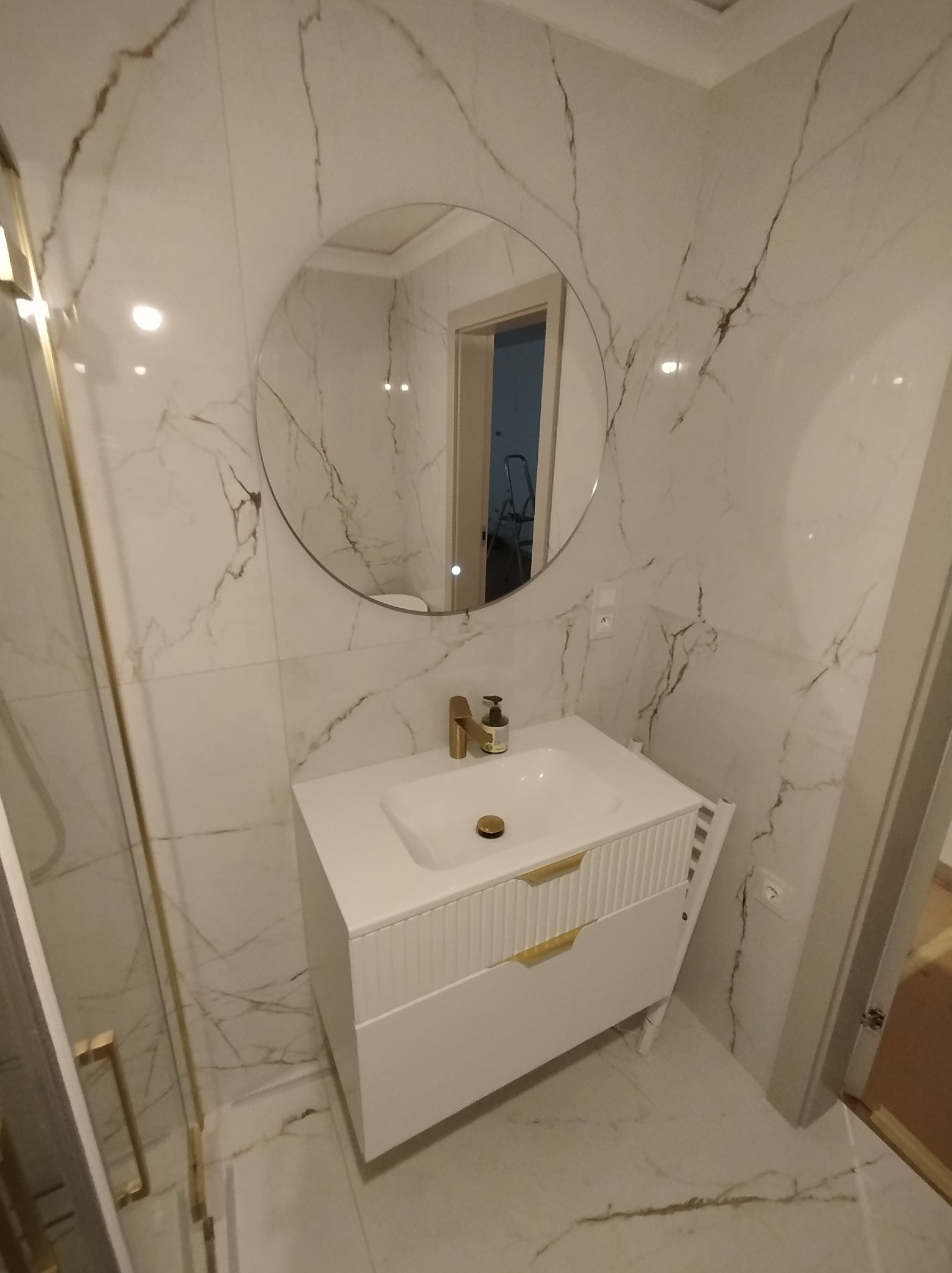 Biała łazienka z marmurowymi płytkami, okrągłym lustrem z podświetleniem i szafką umywalkową z frezowanymi frontami. Złote detale armatury i uchwytów.