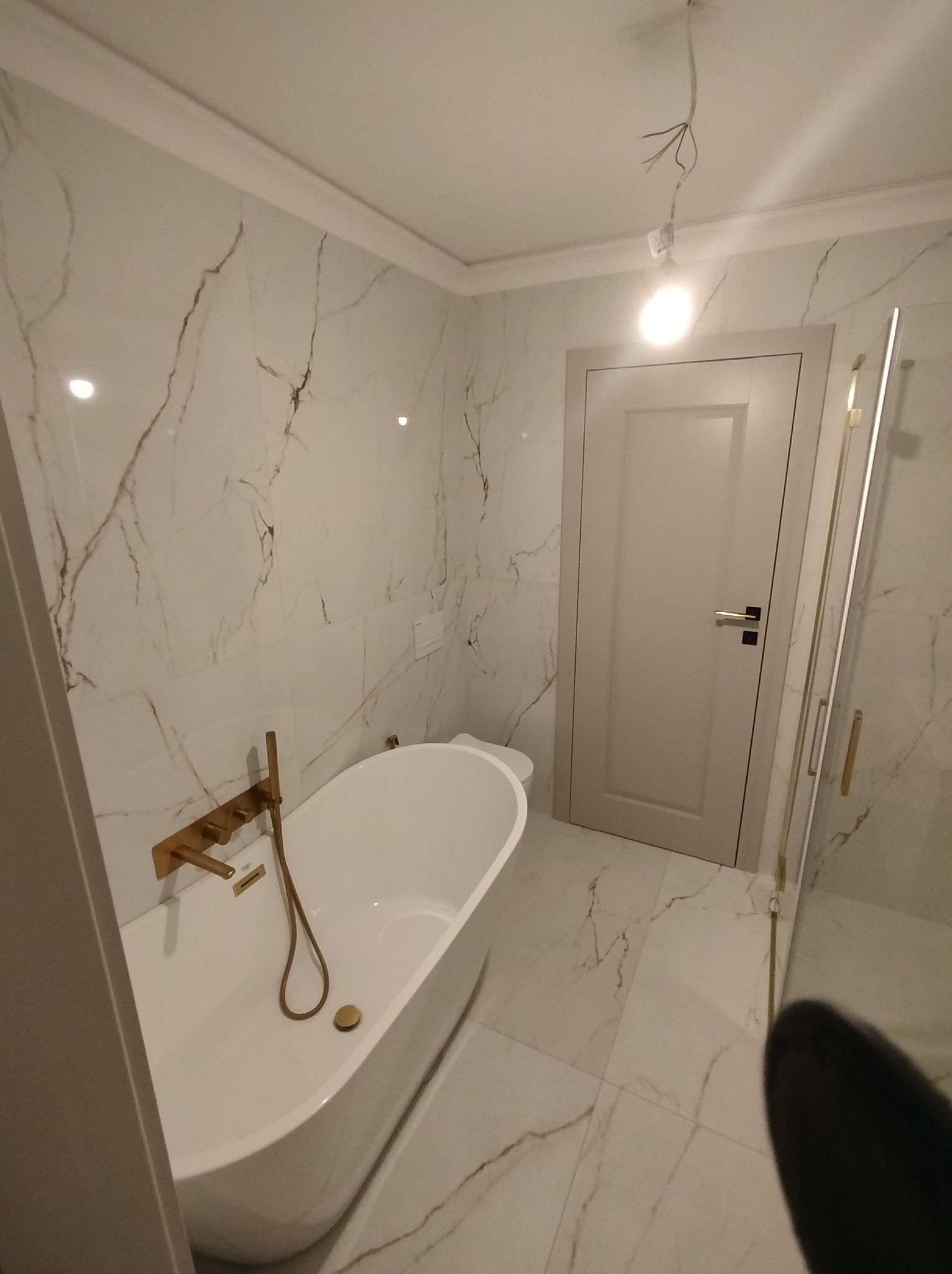 Elegancka łazienka z wanną wolnostojącą i marmurowymi płytkami. Złota armatura, szklana kabina prysznicowa i białe drzwi tworzą spójną całość. Widoczne przewody oświetleniowe.