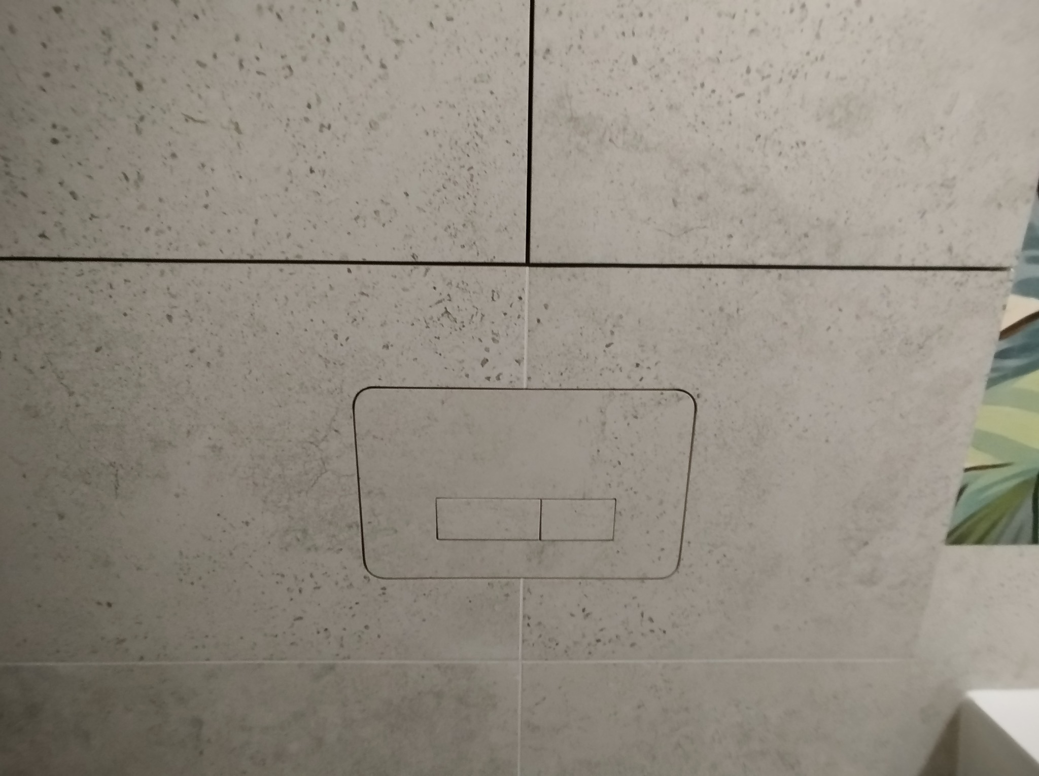 Szare, matowe płytki imitujące kamień z ukrytym przyciskiem spłuczki w łazience. Minimalistyczny design, precyzyjne fugi, fragment roślinnego motywu w tle.