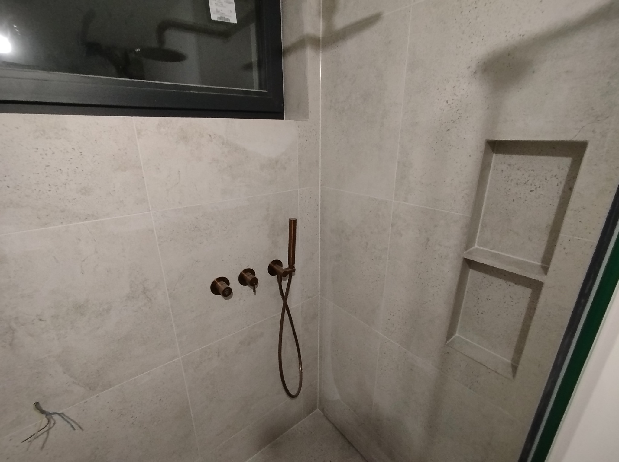 Wykończona kabina prysznicowa z beżowymi płytkami, wnęką na kosmetyki i miedzianą armaturą. Widoczne przewody instalacji. Minimalistyczny design.