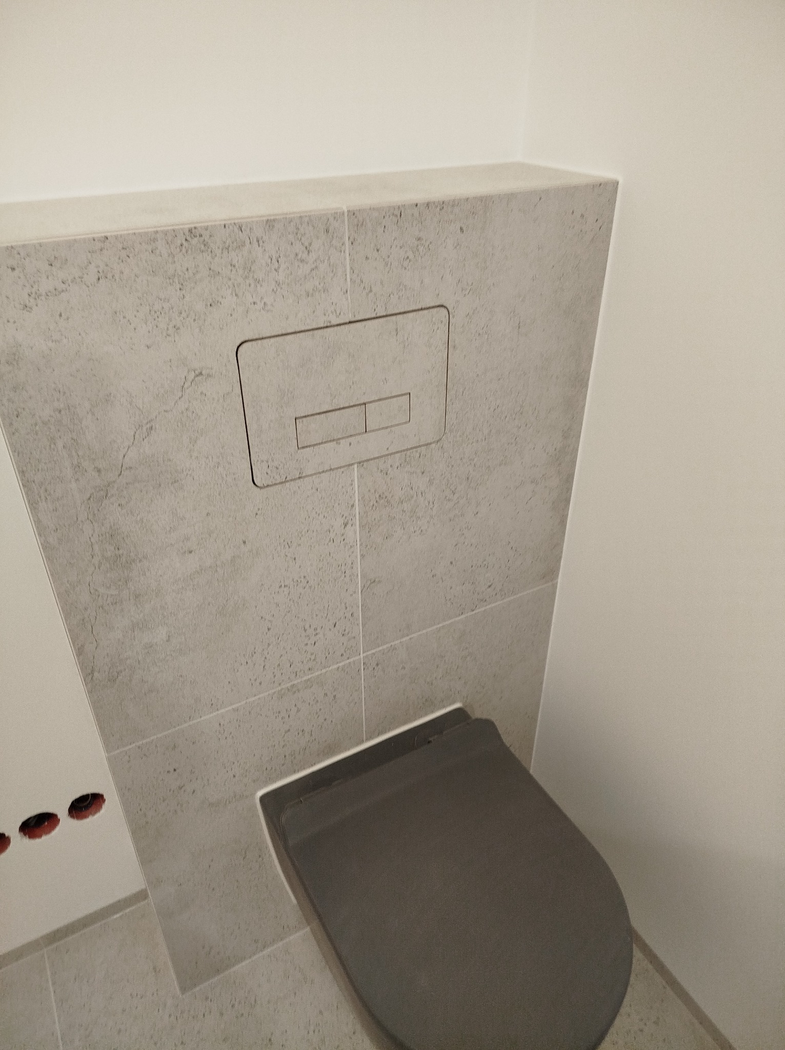 Szary, podtynkowy zestaw toaletowy z płytkami imitującymi beton i ciemnoszarą deską sedesową w minimalistycznej łazience.