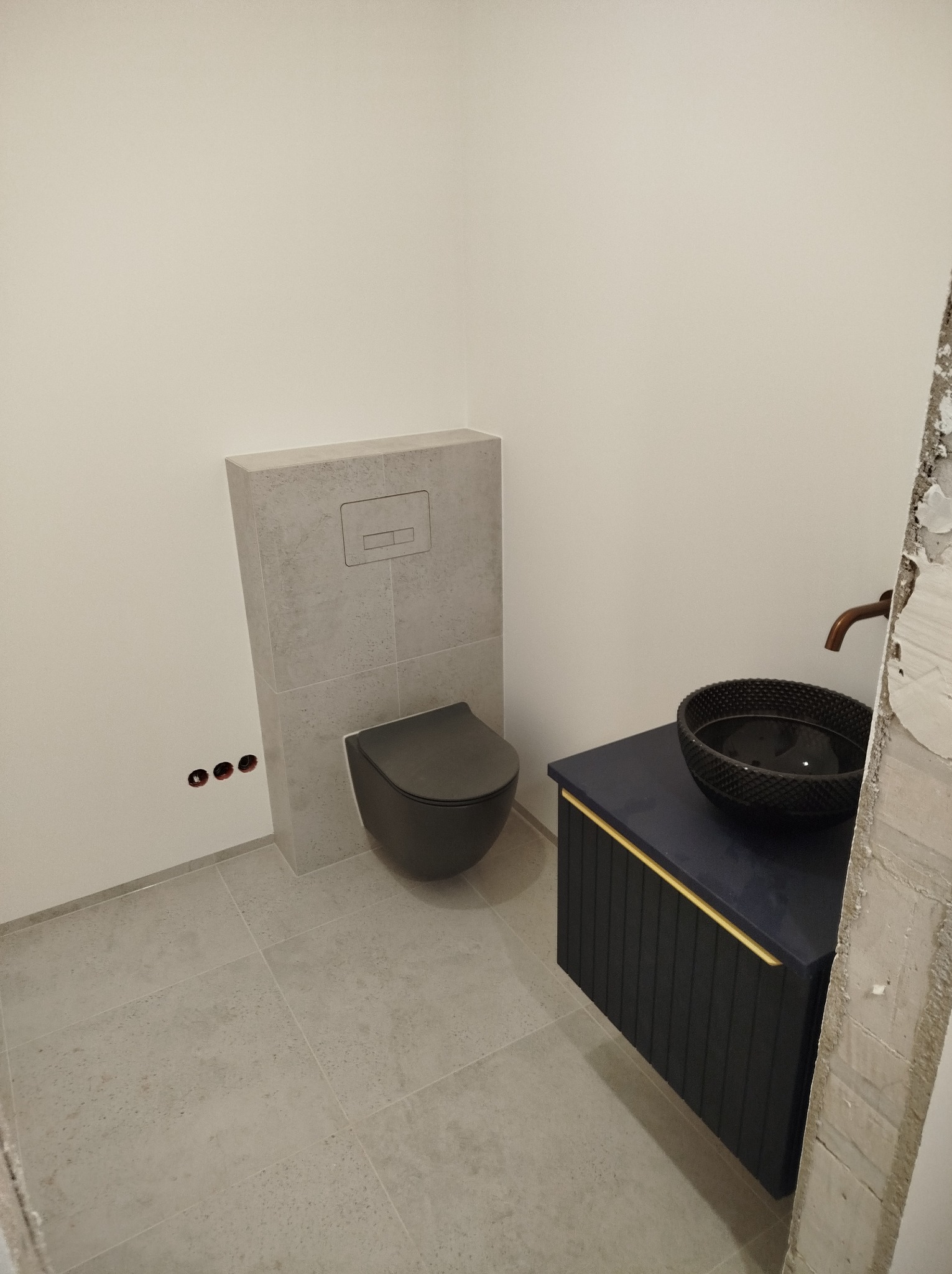 Nowoczesna łazienka z podwieszaną toaletą i umywalką w czarnej misie na szafce w kolorze granatowym, wykończona szarą płytką podłogową i elementami w odcieniach szarości.