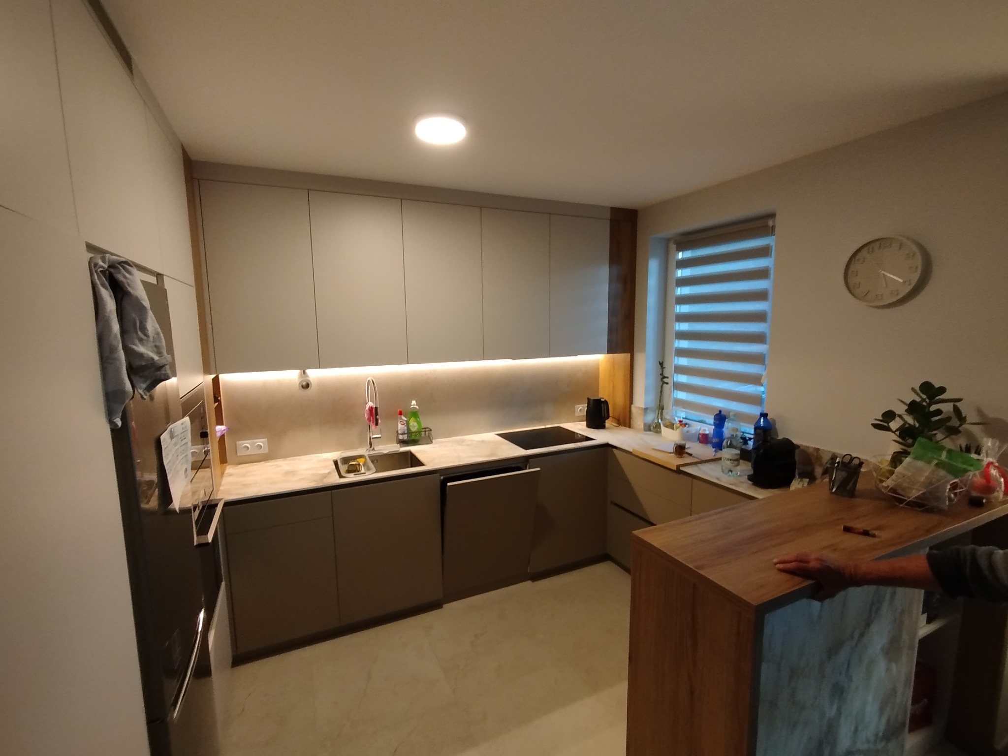 Nowoczesna kuchnia w odcieniach szarości z drewnianym blatem, oświetleniem LED pod szafkami i marmurową podłogą. Minimalistyczny design z zegarem na ścianie.