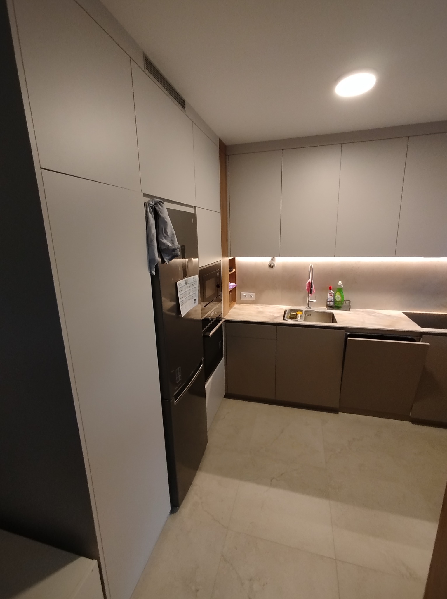 Nowoczesna kuchnia w odcieniach szarości z marmurową podłogą, oświetleniem LED pod szafkami i zabudowaną lodówką z notatkami na drzwiach.