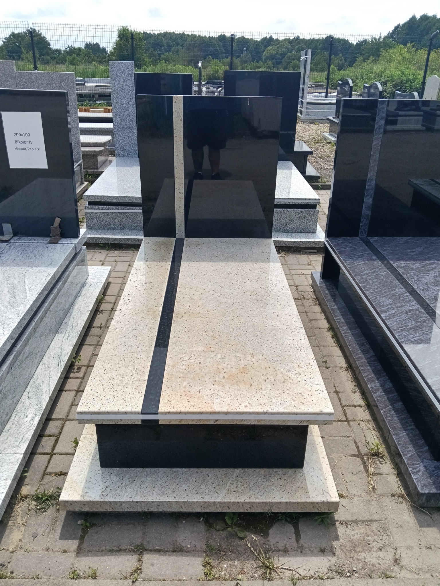 Nowoczesny nagrobek z polerowanego czarnego i jasnego granitu, z wyraźnym geometrycznym wzorem, eksponowany na zewnątrz w otoczeniu innych pomników.