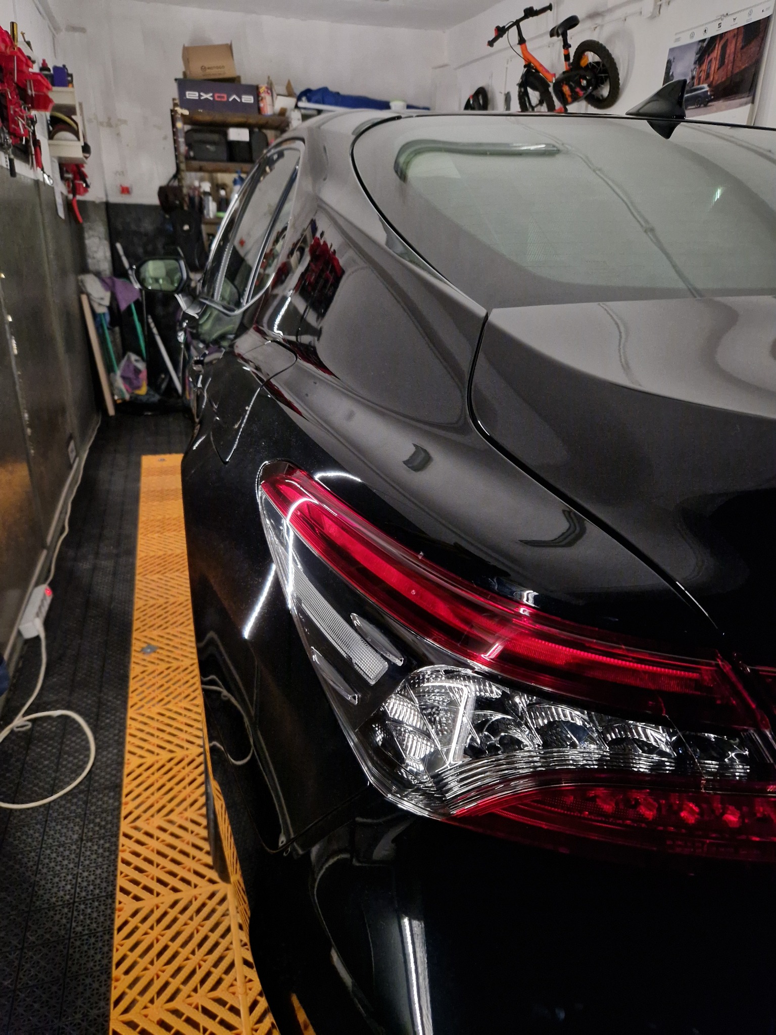 Błyszczący, czarny samochód w garażu, widoczny tylny reflektor z chromowanymi elementami i czerwonymi światłami, podłoga z żółtych, ażurowych paneli.