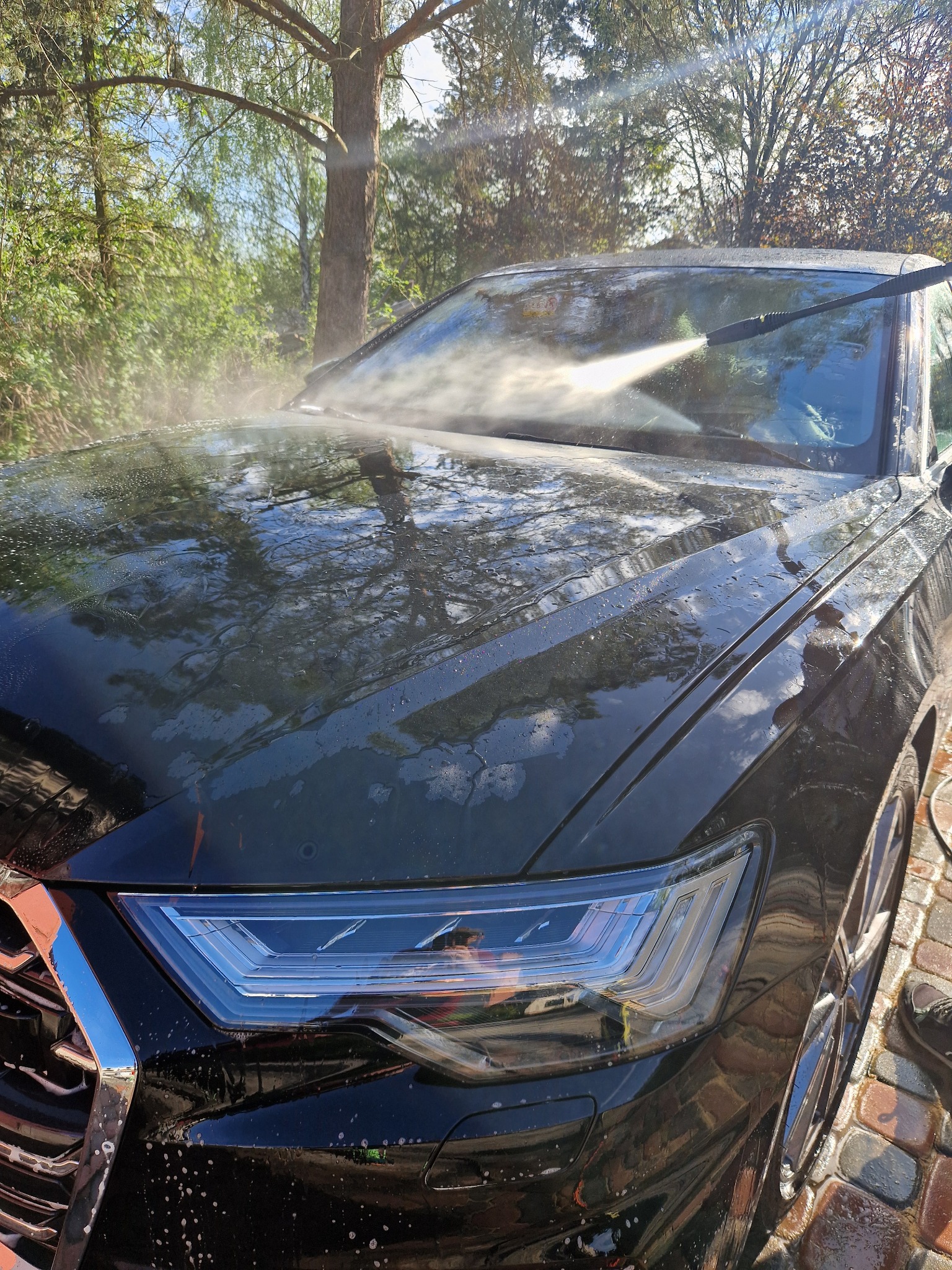 Czarny samochód osobowy myty myjką ciśnieniową na zewnątrz, z widocznymi kroplami wody na karoserii i refleksami drzew w lakierze.