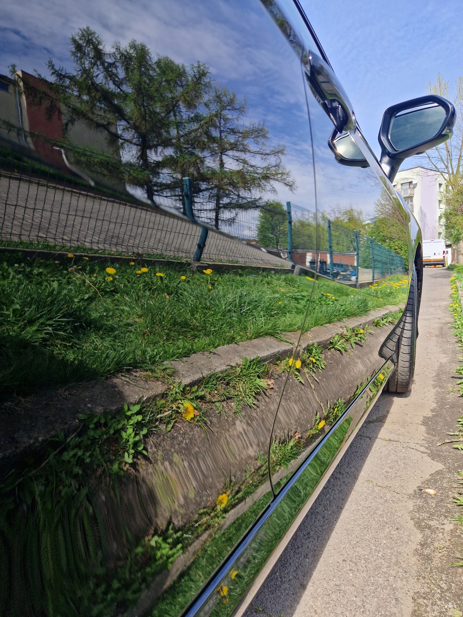 Błyszcząca karoseria czarnego samochodu odbija krajobraz z trawnikiem, betonowym krawężnikiem i budynkiem w tle.