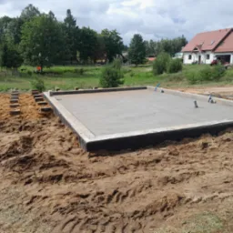 Wylany betonowy fundament płyty pod budowę domu jednorodzinnego, widoczne szalunki i wystające rury instalacyjne, teren wyrównany, w tle dom w stanie surowym otwartym.