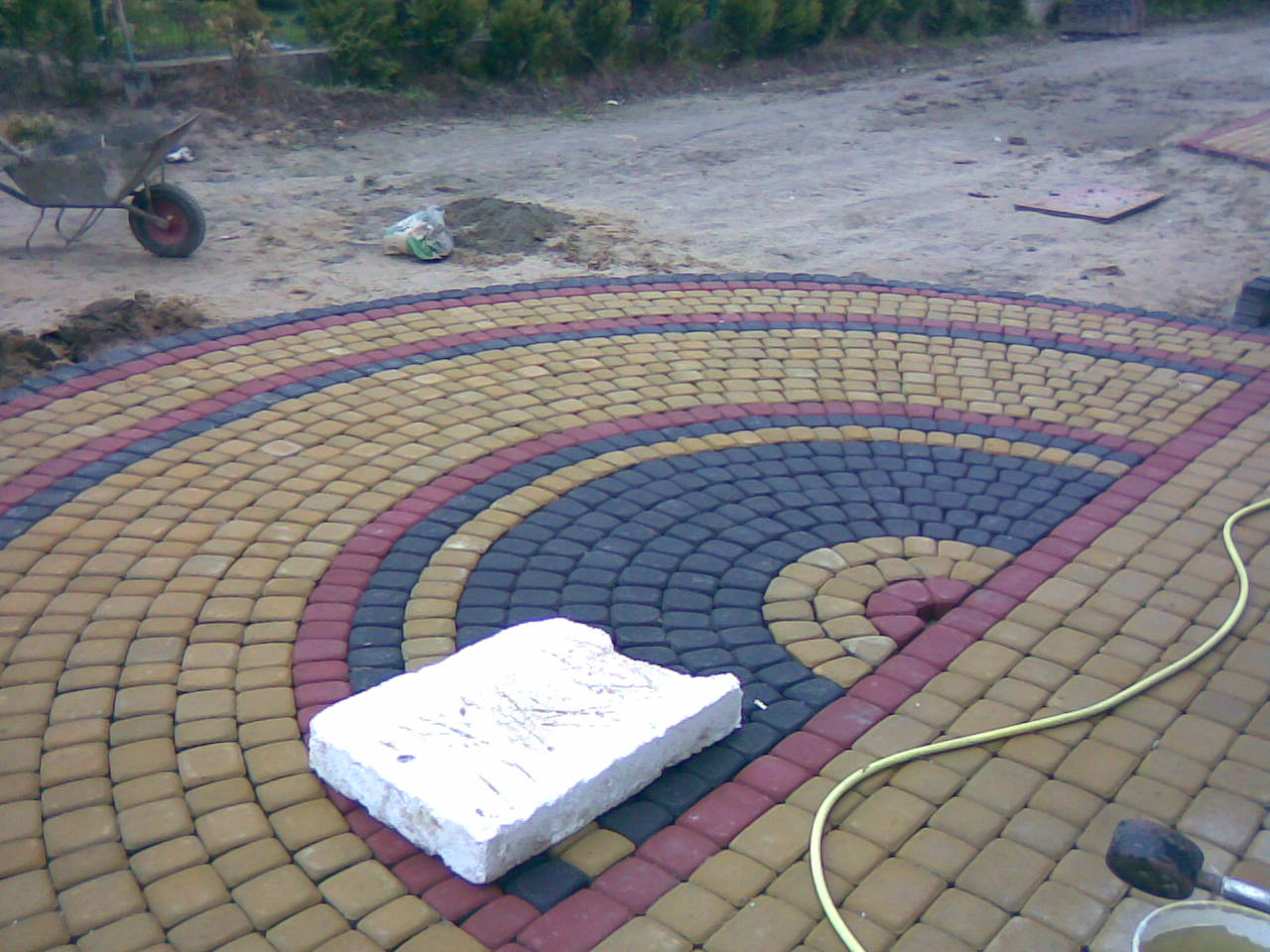 Okładzina z kostki brukowej ułożona w koncentryczne kręgi z kolorowych elementów, z widocznym fragmentem węża ogrodowego i niedokończonym obszarem z ziemią, taczka w tle.