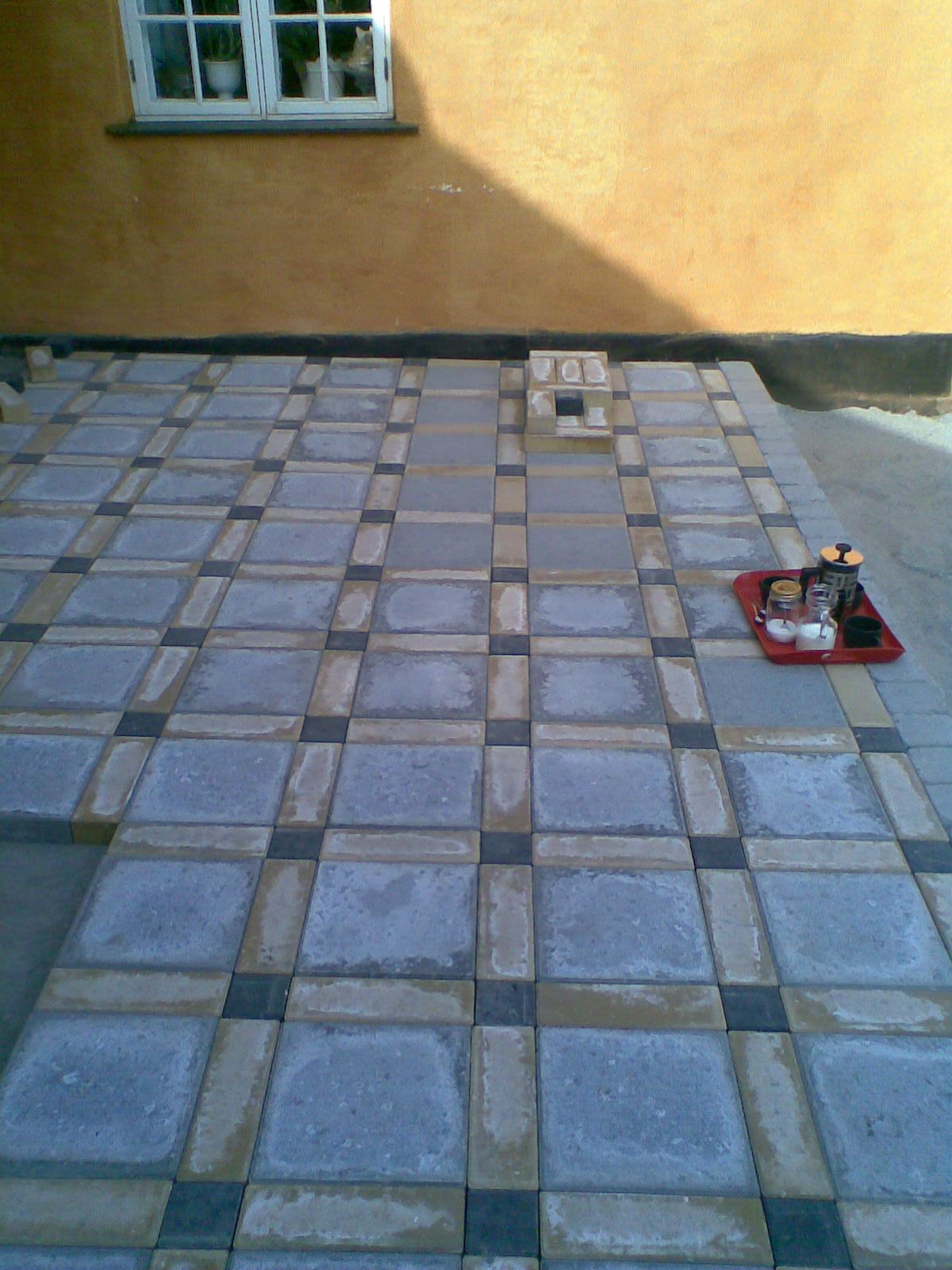 Niedokończony taras z kostki brukowej w odcieniach szarości, beżu i czerni, z elementami dekoracyjnymi ułożonymi w formie miniaturowego kominka i tacą z kawą. W tle żółta ściana budynku z oknem,...