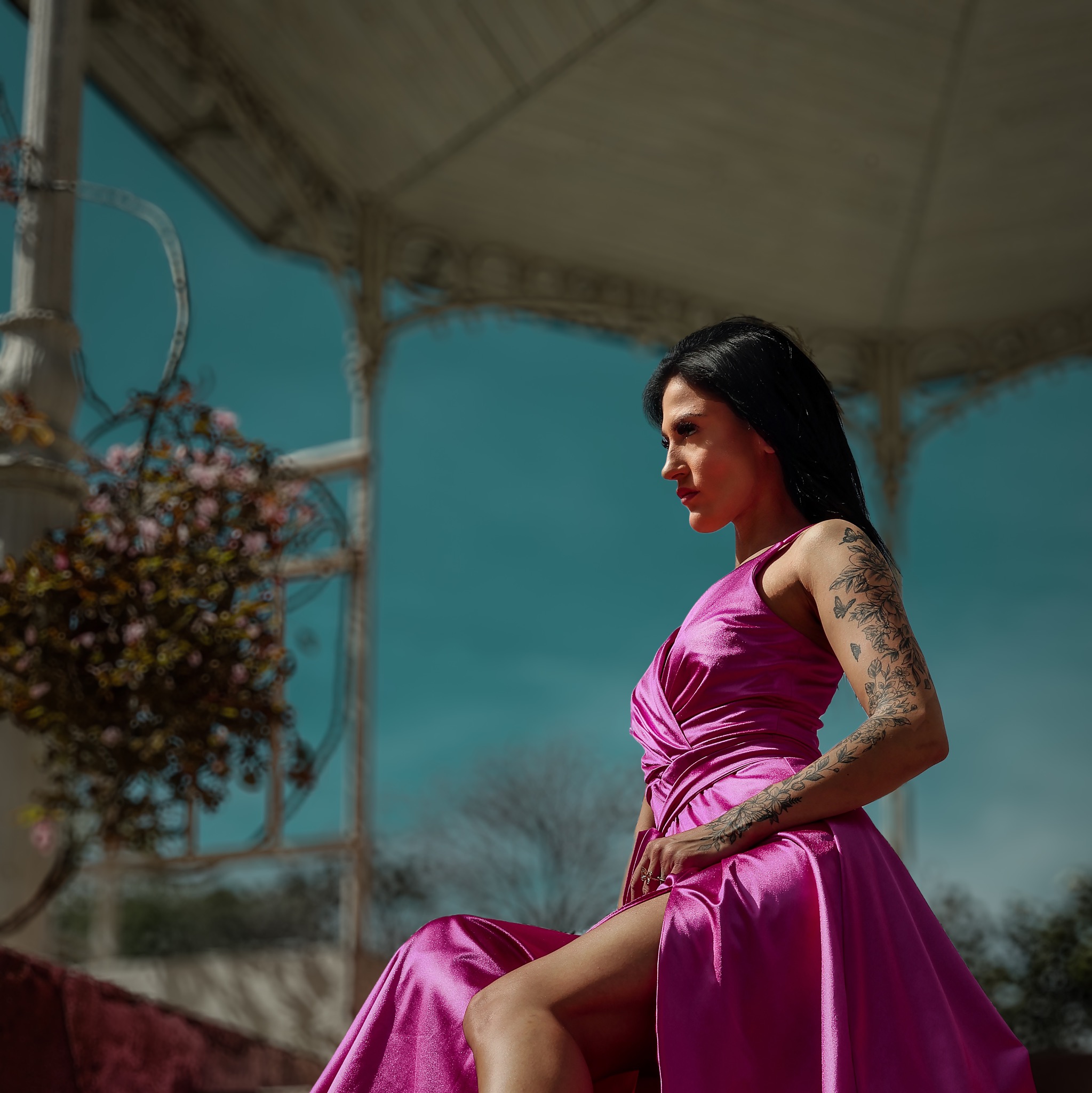 Kobieta w błyszczącej, fuksjowej sukni z rozcięciem siedzi na tle ażurowej altany z kwitnącymi pnączami, eksponując tatuaż na ramieniu.
