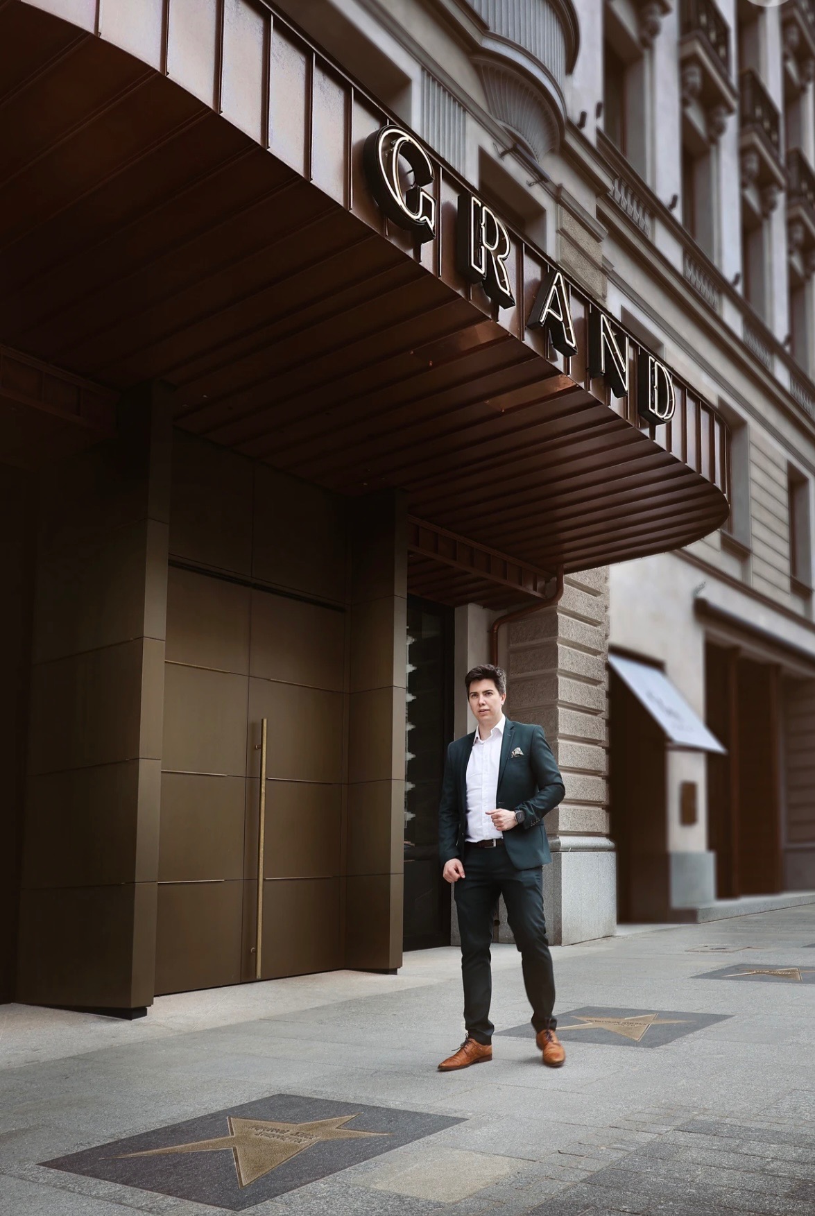 Mężczyzna w ciemnozielonym garniturze pozuje na tle budynku z napisem 'GRAND' nad wejściem, na chodniku gwiazdy w stylu Alei Gwiazd.