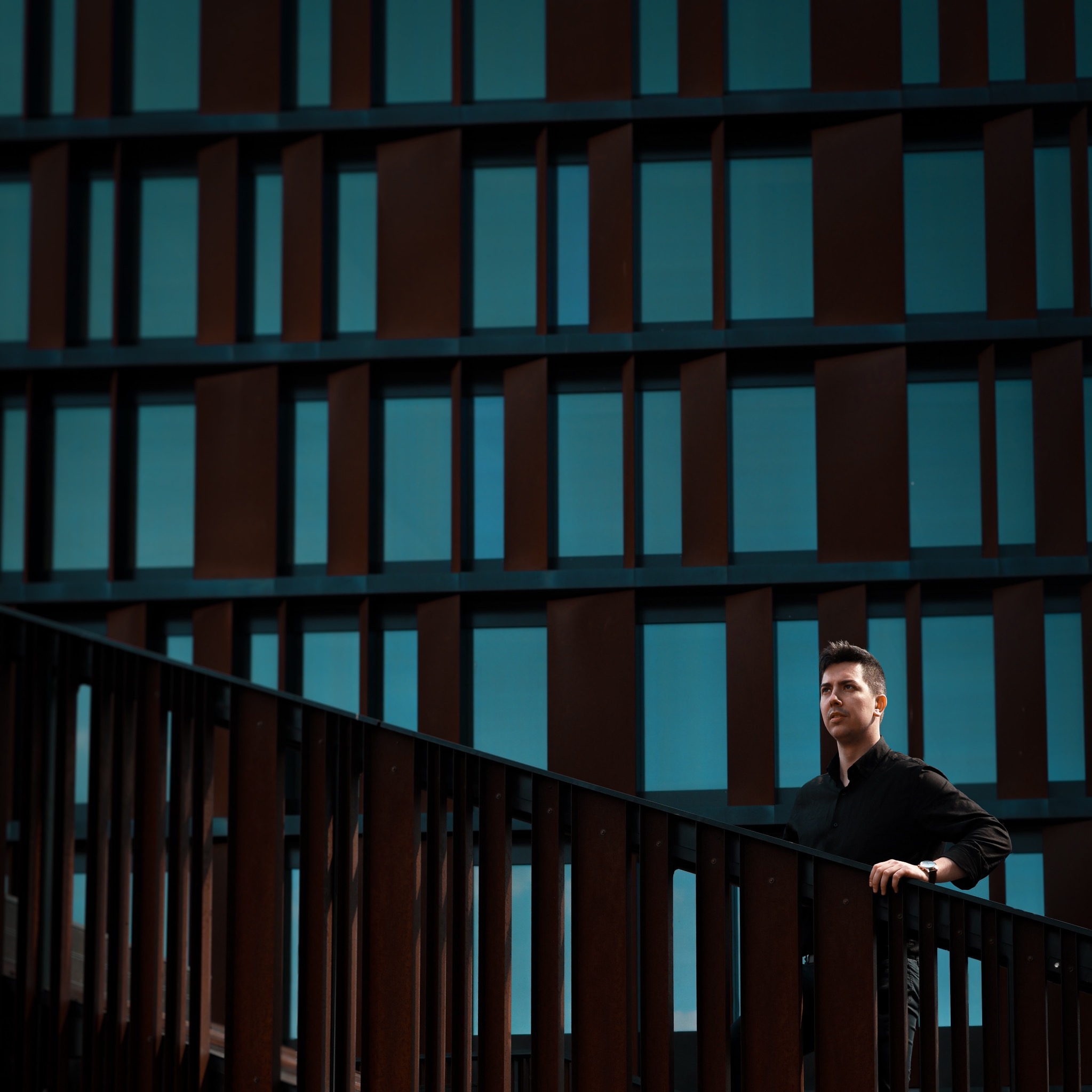 Mężczyzna w czarnej koszuli opiera się o metalową barierkę na tle nowoczesnego budynku z rustykalnymi panelami okiennymi.