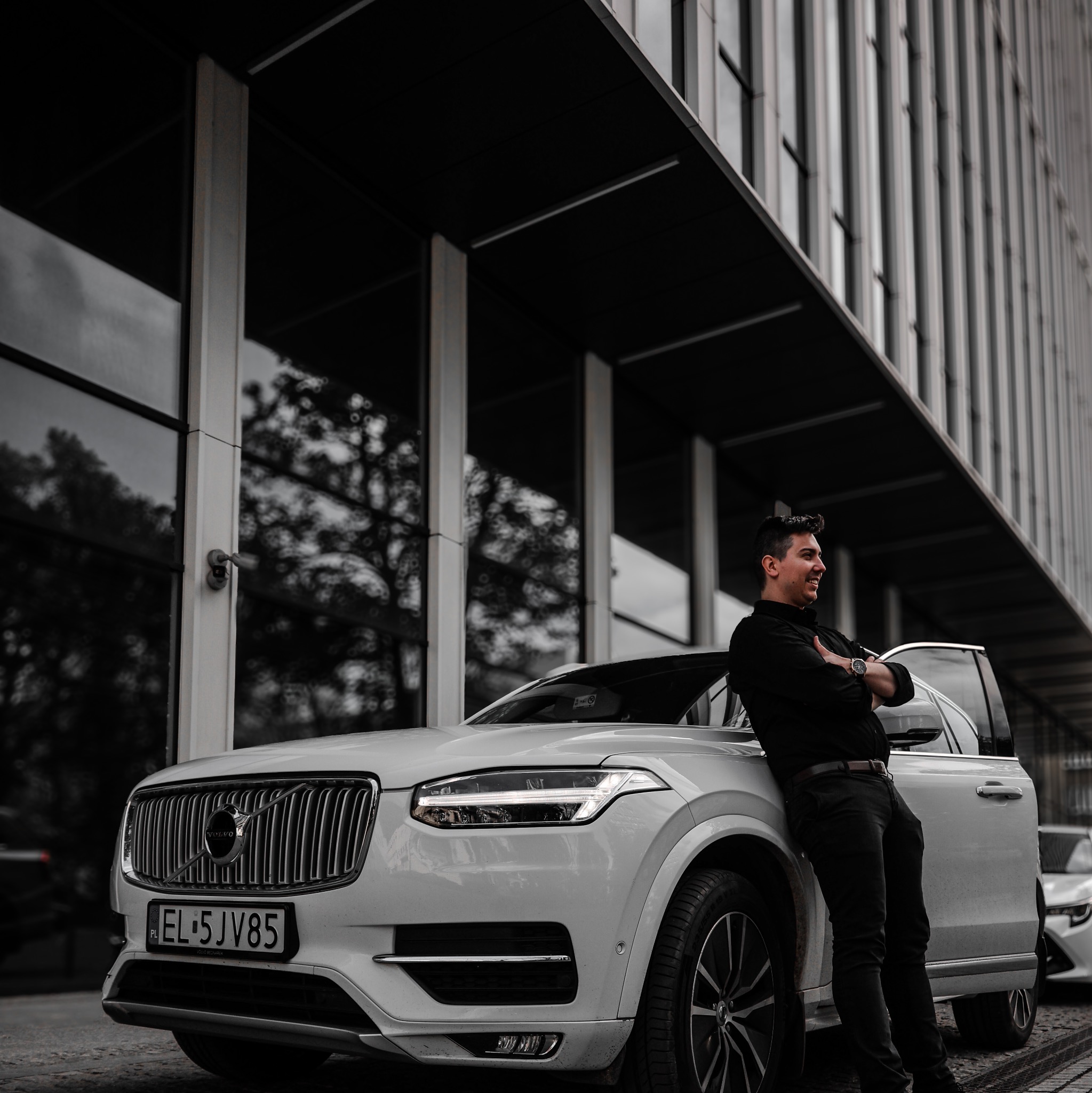 Uśmiechnięty mężczyzna w czarnym ubraniu opiera się o białe Volvo XC90 na tle nowoczesnego budynku biurowego z szklaną fasadą.