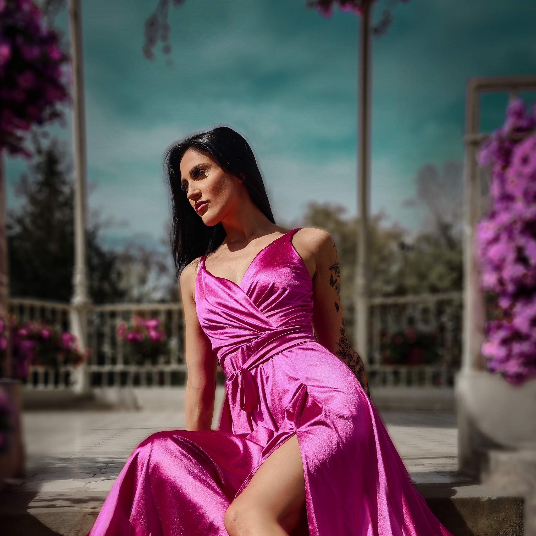Kobieta w satynowej sukni w kolorze fuksji siedzi w altanie z kwiatami, patrząc w bok, z tatuażem na ramieniu.