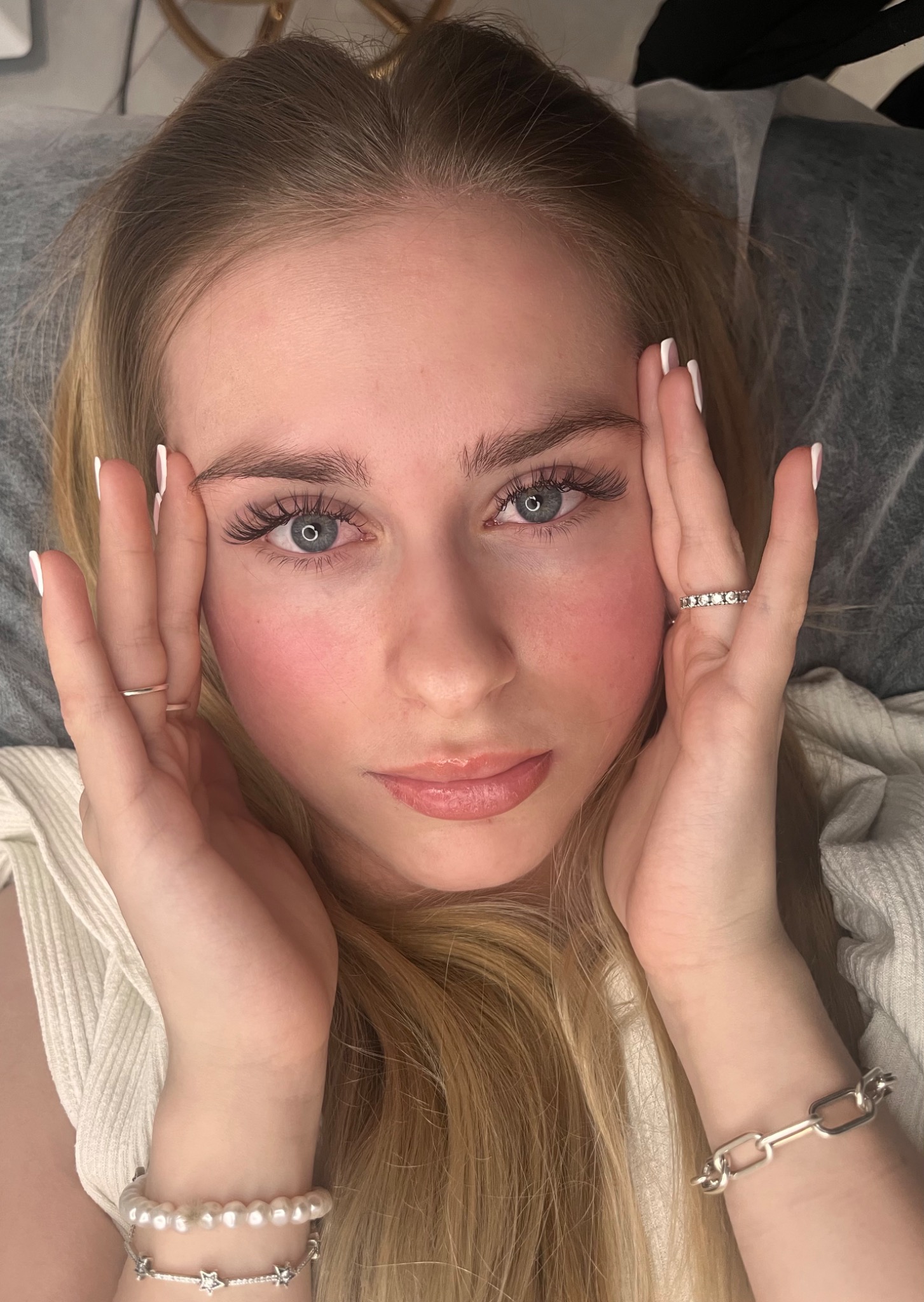 Portret młodej kobiety z przedłużonymi rzęsami, dłonie z białym manicure przy twarzy, widoczne pierścionki i bransoletki, ujęcie z góry.