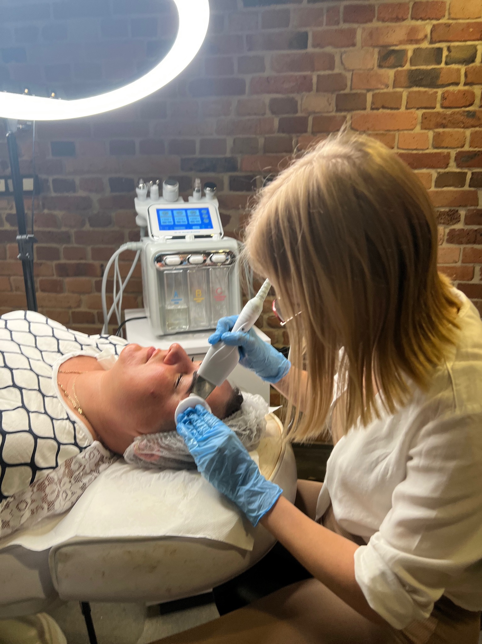 Klientka podczas zabiegu oczyszczania wodorowego twarzy w gabinecie kosmetycznym; widoczna maszyna do zabiegów i lampa pierścieniowa.