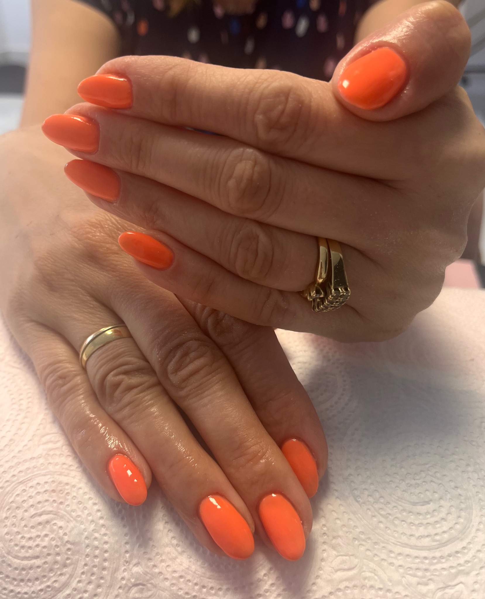 Dłonie z pomarańczowym manicure hybrydowym o migdałowym kształcie, widoczny złoty pierścionek z cyrkoniami na jednej dłoni i obrączka na drugiej, na podkładzie z papieru.