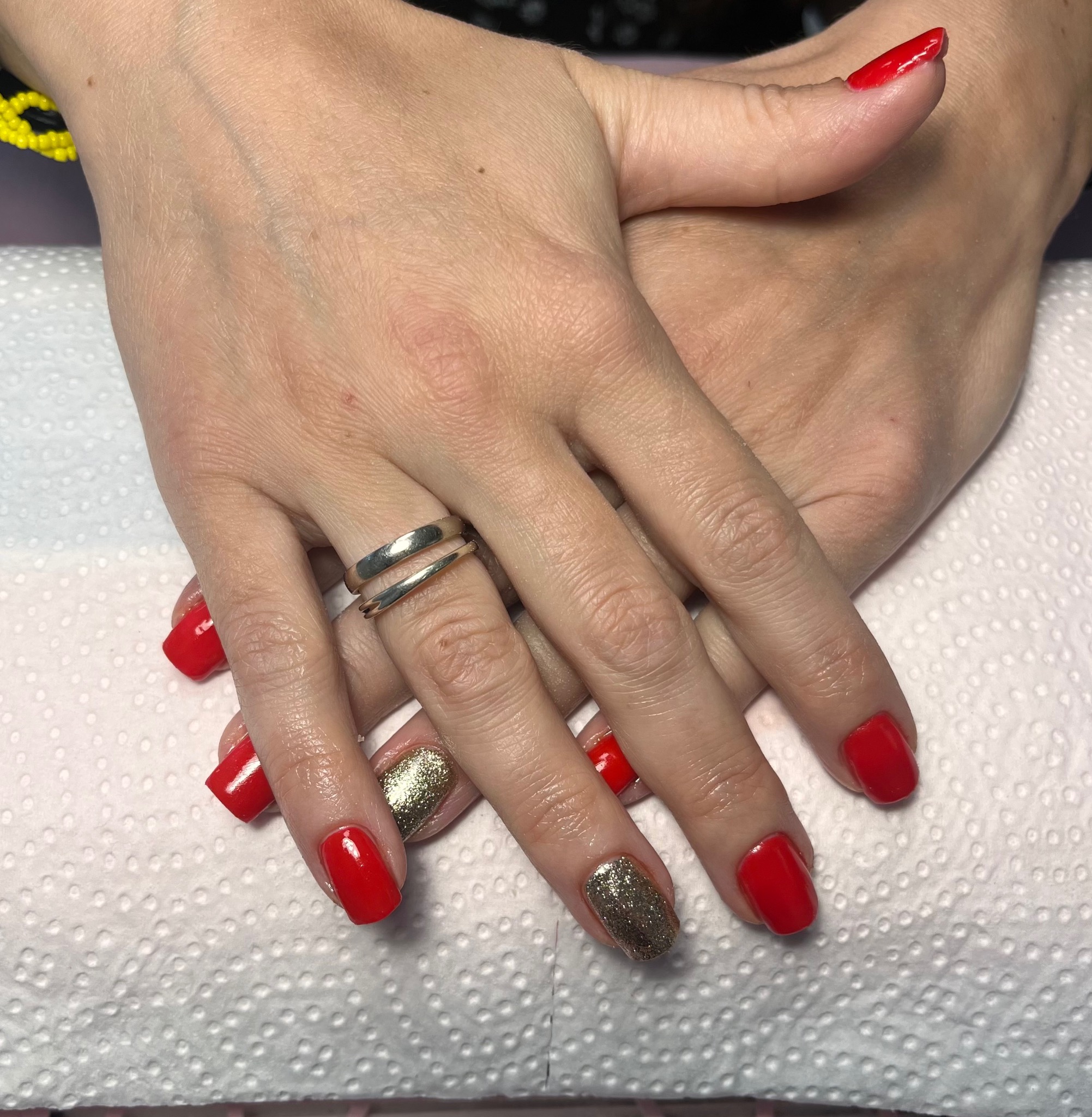 Dłonie kobiety z czerwonym i złotym brokatowym manicure, na serdecznym palcu srebrny pierścionek z dwoma obręczami, na białym podkładzie.