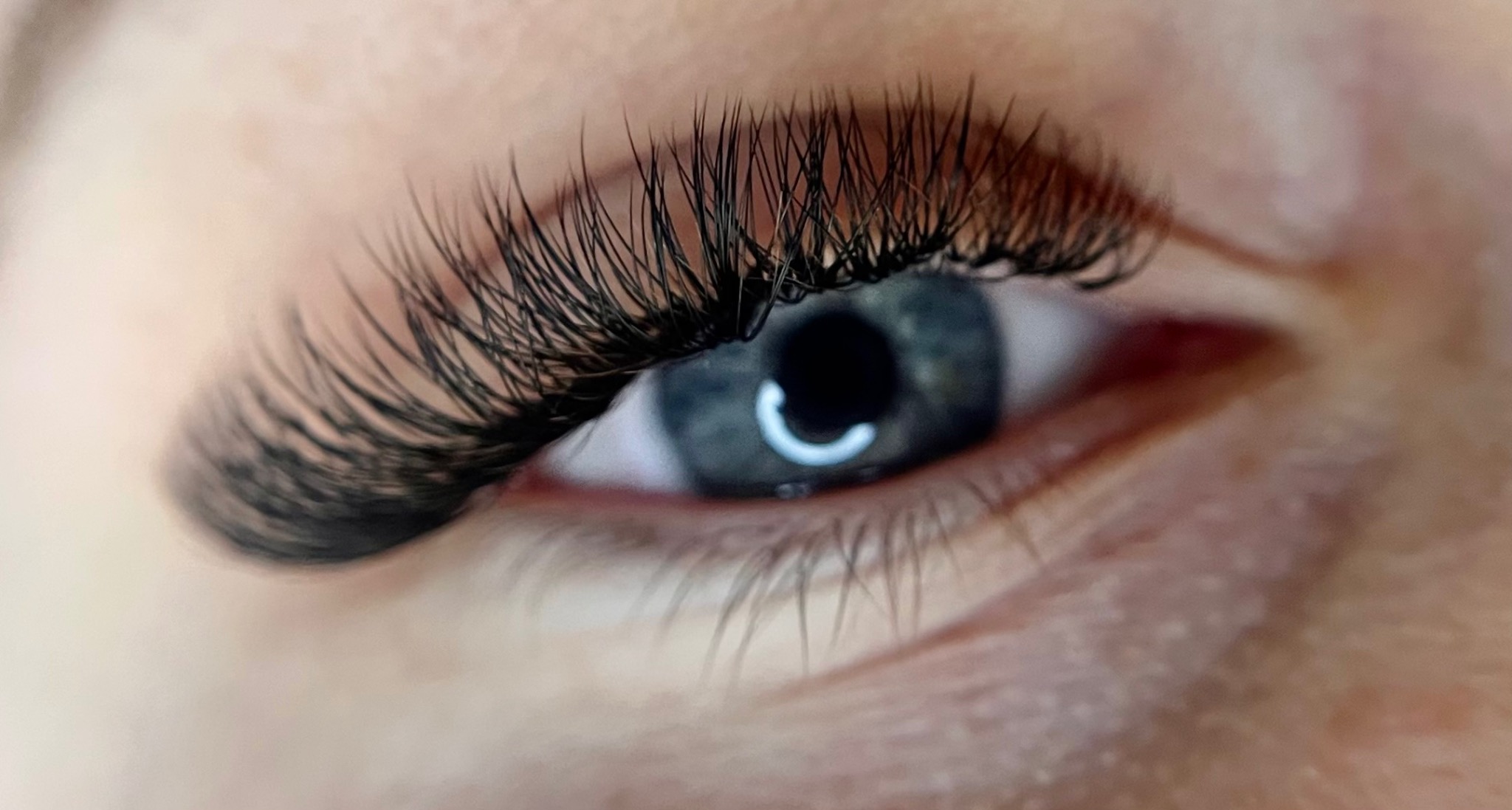 Zbliżenie na oko z gęstymi, profesjonalnie przedłużonymi czarnymi rzęsami, podkreślającymi naturalny, szaro-niebieski kolor tęczówki. Widoczne delikatne zmarszczki wokół oka.