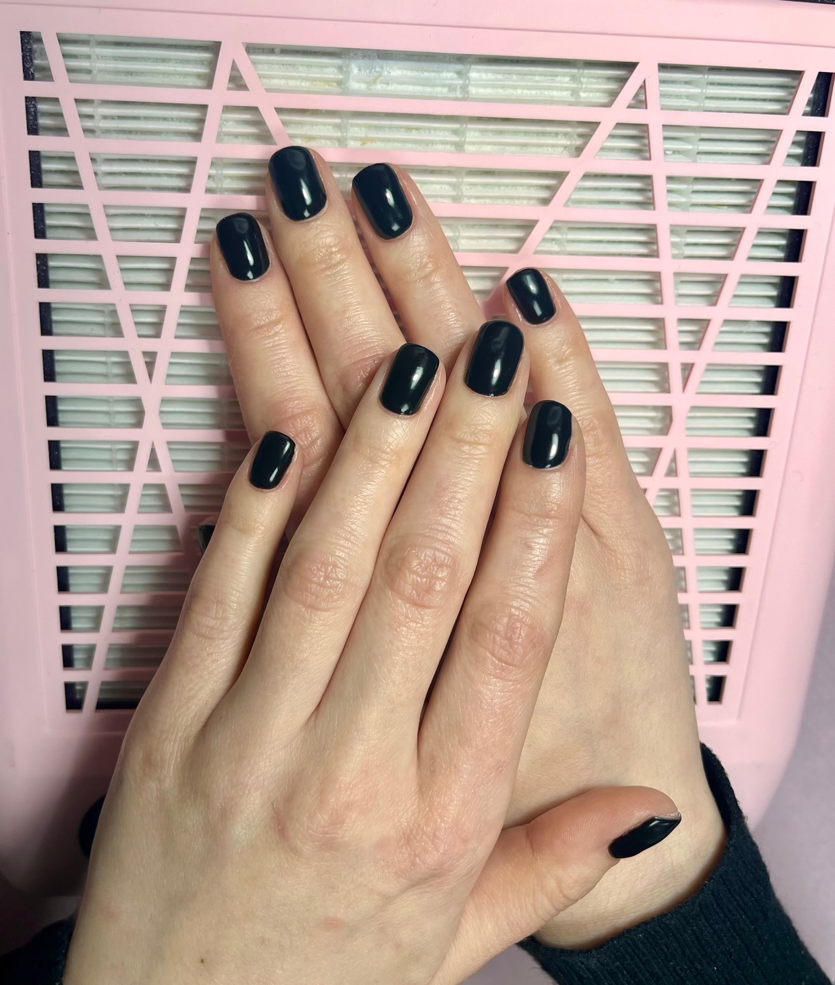 Dłonie z świeżo pomalowanymi, błyszczącymi, czarnymi paznokciami na tle różowego pochłaniacza pyłu do paznokci.