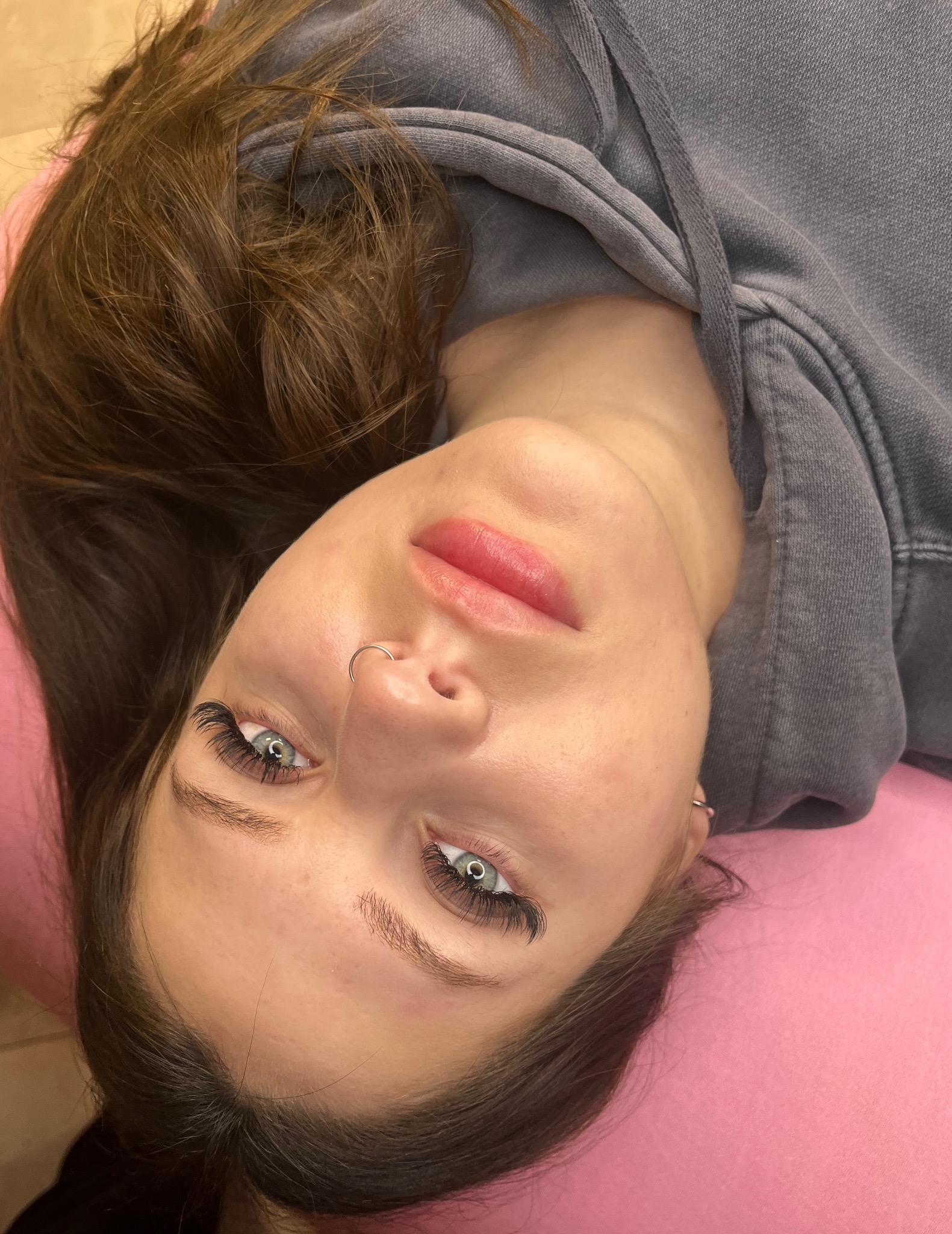Zbliżenie na twarz kobiety z widocznymi, profesjonalnie przedłużonymi rzęsami i delikatnym makijażem ust. Piercing w nosie. Widoczne brązowe włosy i fragment szarej bluzy z kapturem na różowym tle.
