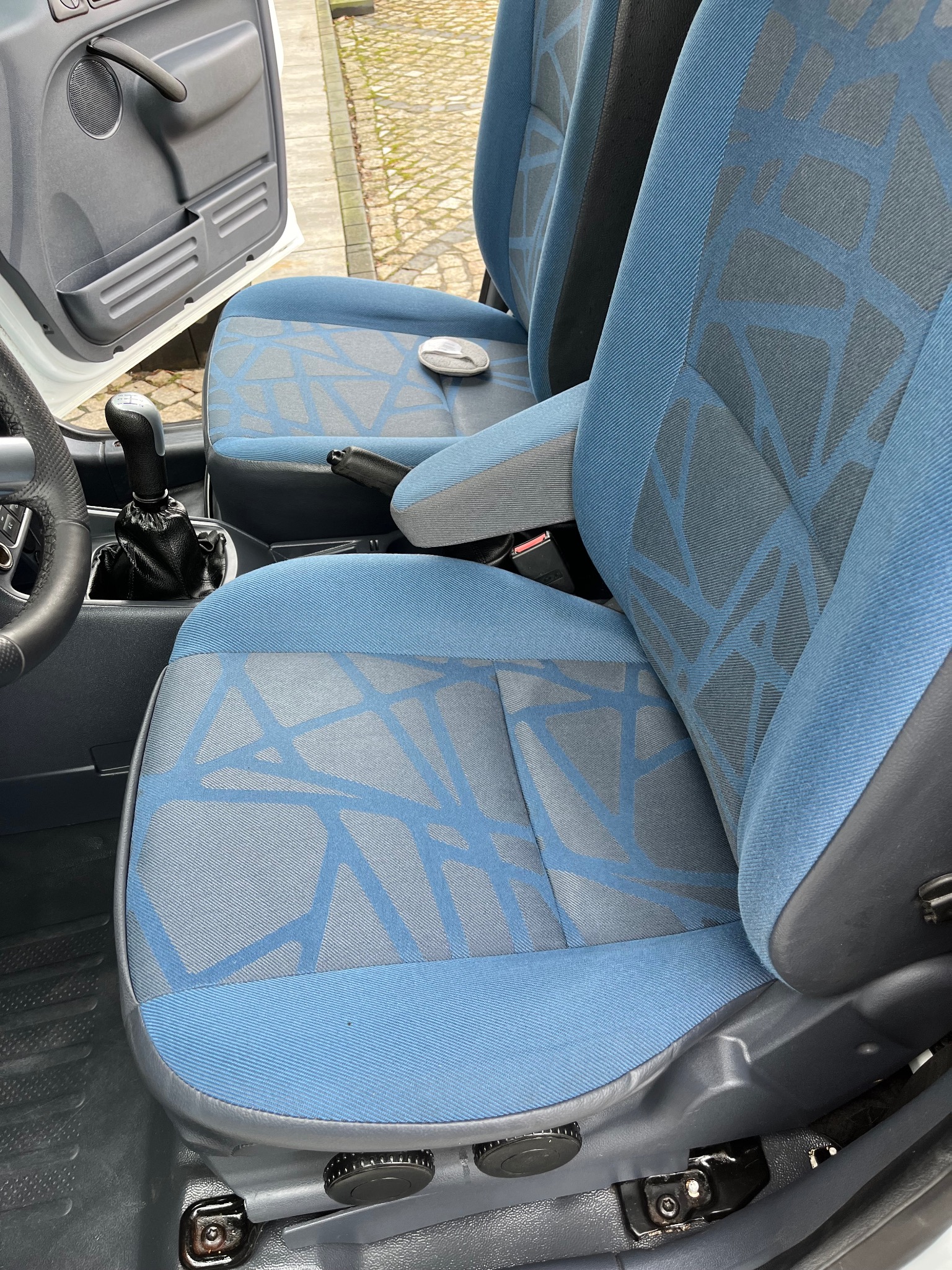 Wnętrze samochodu dostawczego: zbliżenie na błękitno-szare fotele z geometrycznym wzorem, dźwignia zmiany biegów i fragment otwartych drzwi.