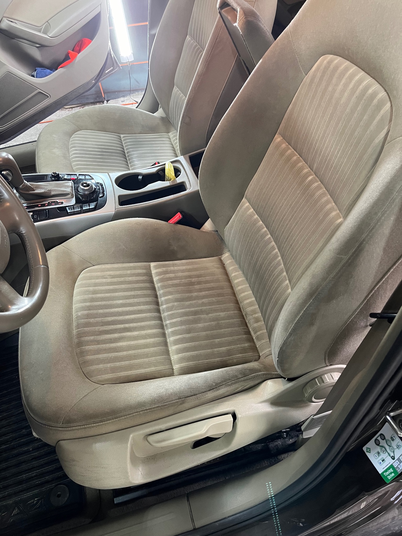 Beżowe, materiałowe fotele samochodowe z widocznymi śladami użytkowania i delikatnymi plamami, wnętrze pojazdu z elementami deski rozdzielczej i kierownicy.